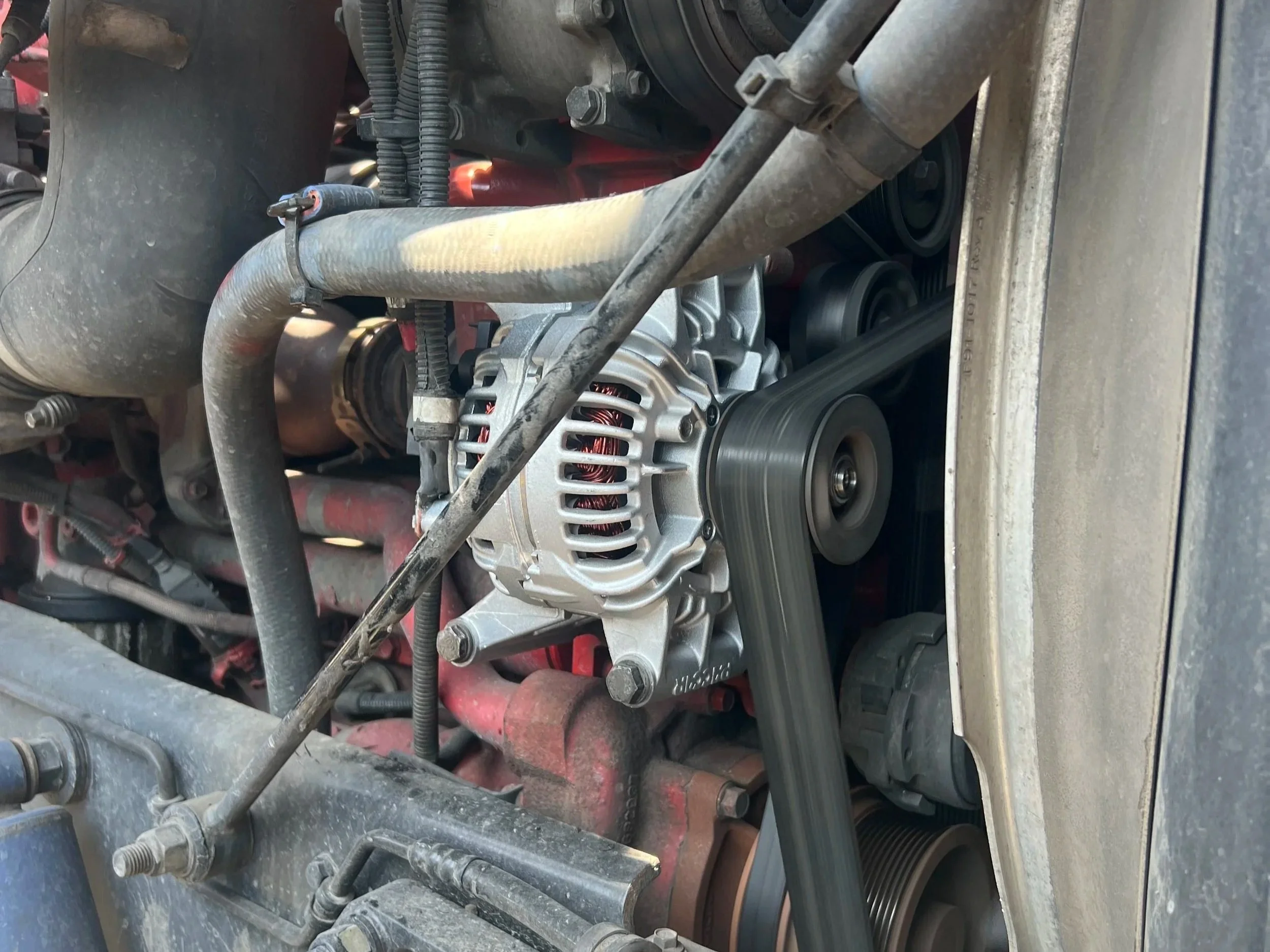 Close-up of a vehicle engine showing a silver alternator with copper wiring, various belts, hoses, and metal components.