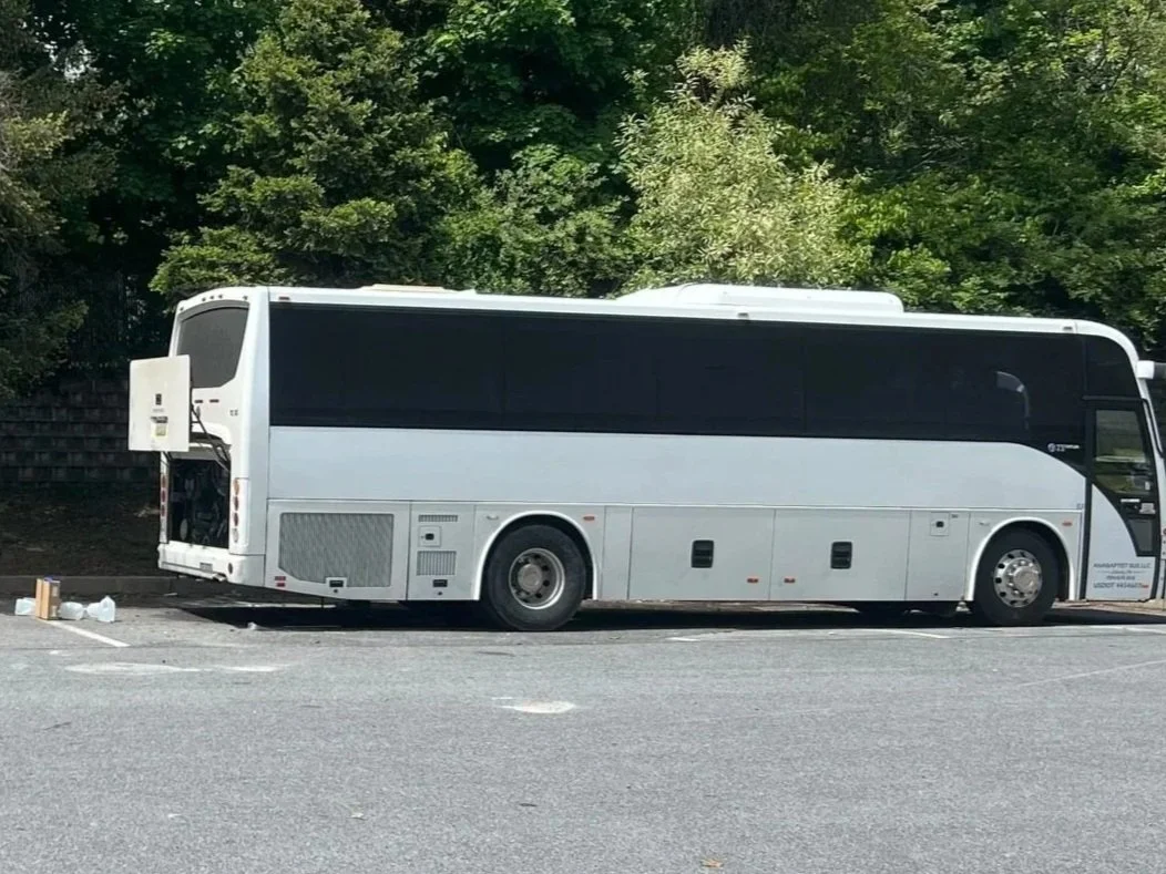 A large white bus with black tinted windows parked in a parking lot, with trees in the background.