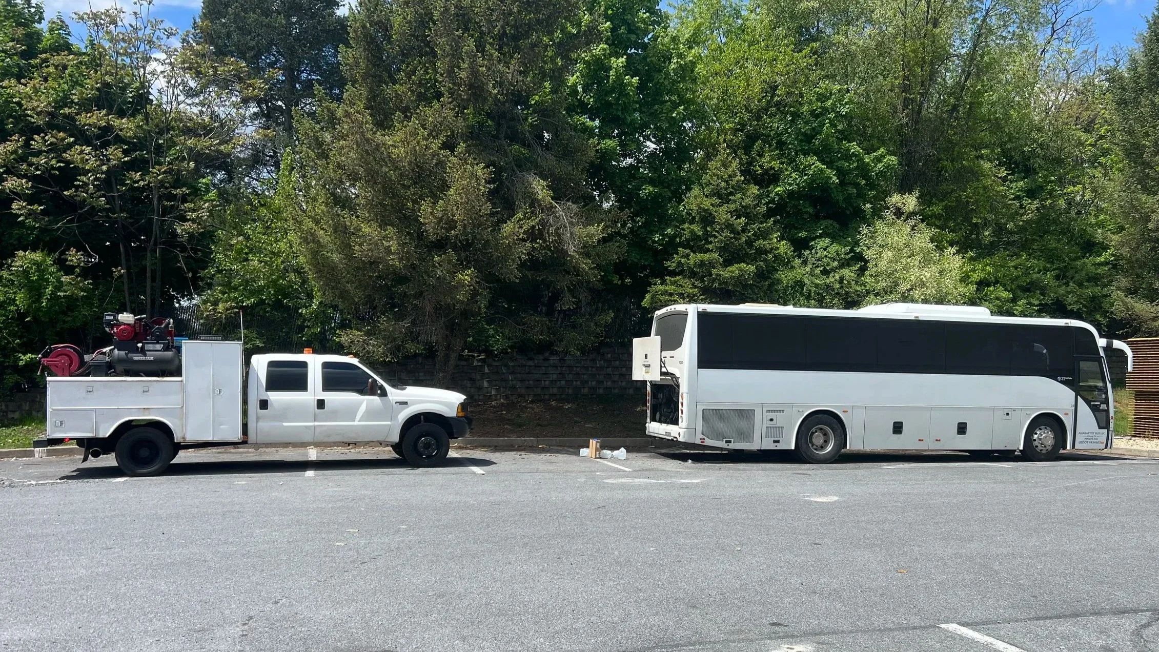 A white utility truck with equipment on its bed and a large white bus parked next to each other in a parking lot, with green trees in the background.