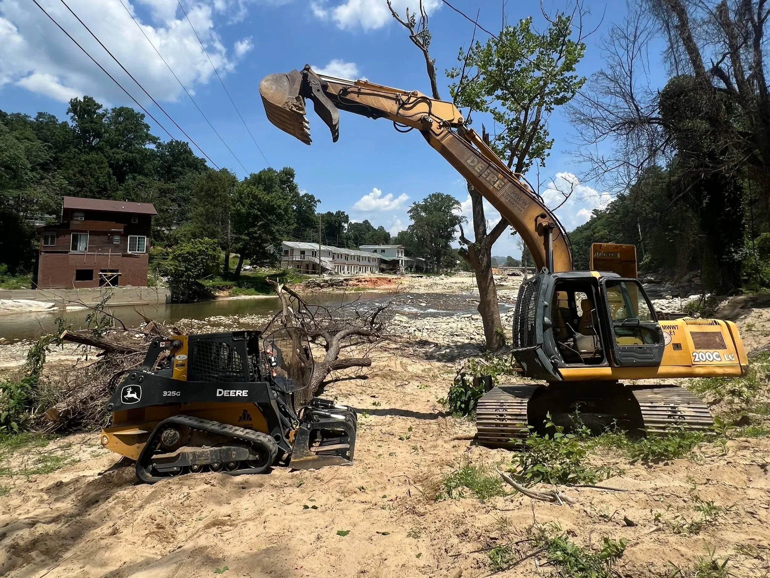 Construction equipment, including excavators and skid steer loader, working on clearing trees and debris near a river, with residential buildings and trees in the background under a partly cloudy sky.
