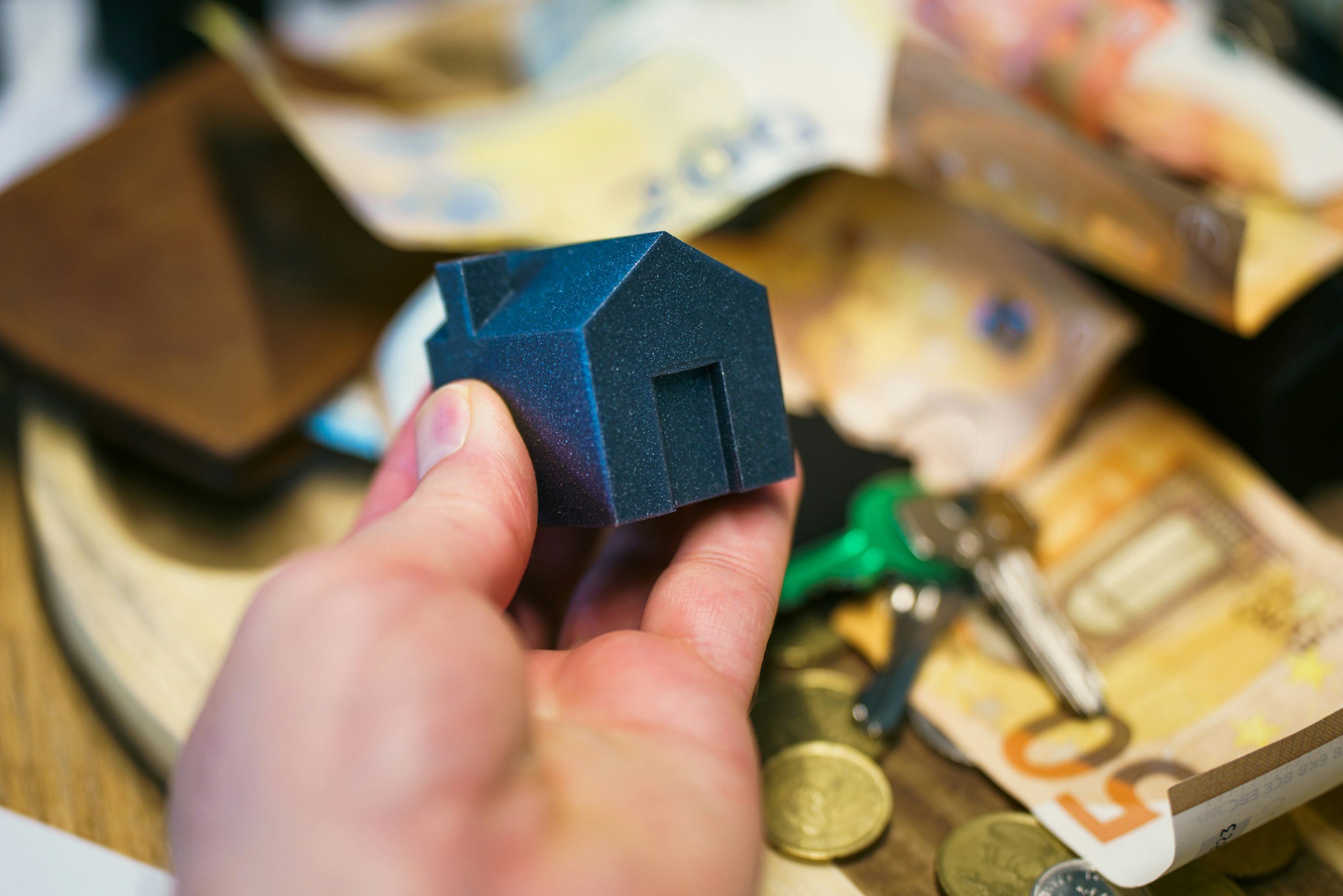 Person holding a small black house-shaped object with a door cutout, surrounded by euros, coins, and keys on a table.