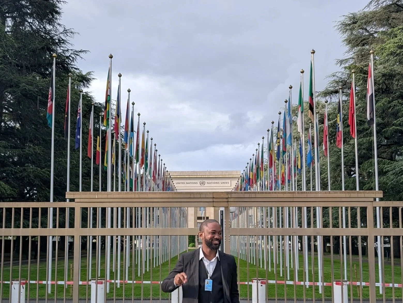 A man in a suit and tie smiling outside the United Nations headquarters, with numerous flagpoles and flags in the background, and a metal gate in the foreground.