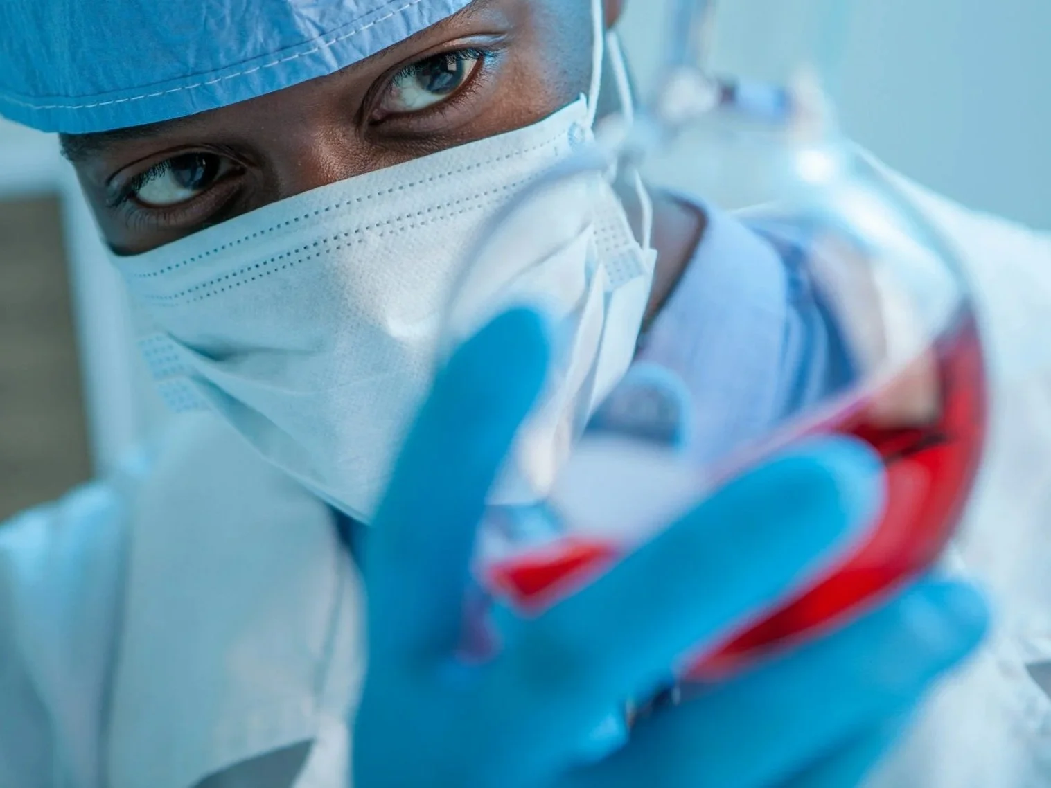 A medical professional in scrubs and a mask holding a petri dish with red liquid, looking directly at the camera.