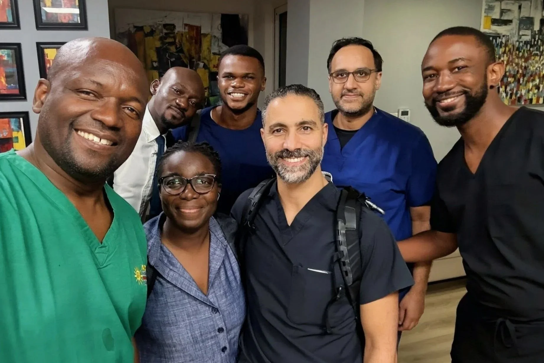 A group of seven diverse healthcare professionals smiling for a selfie indoors, with framed artwork on the walls behind them.