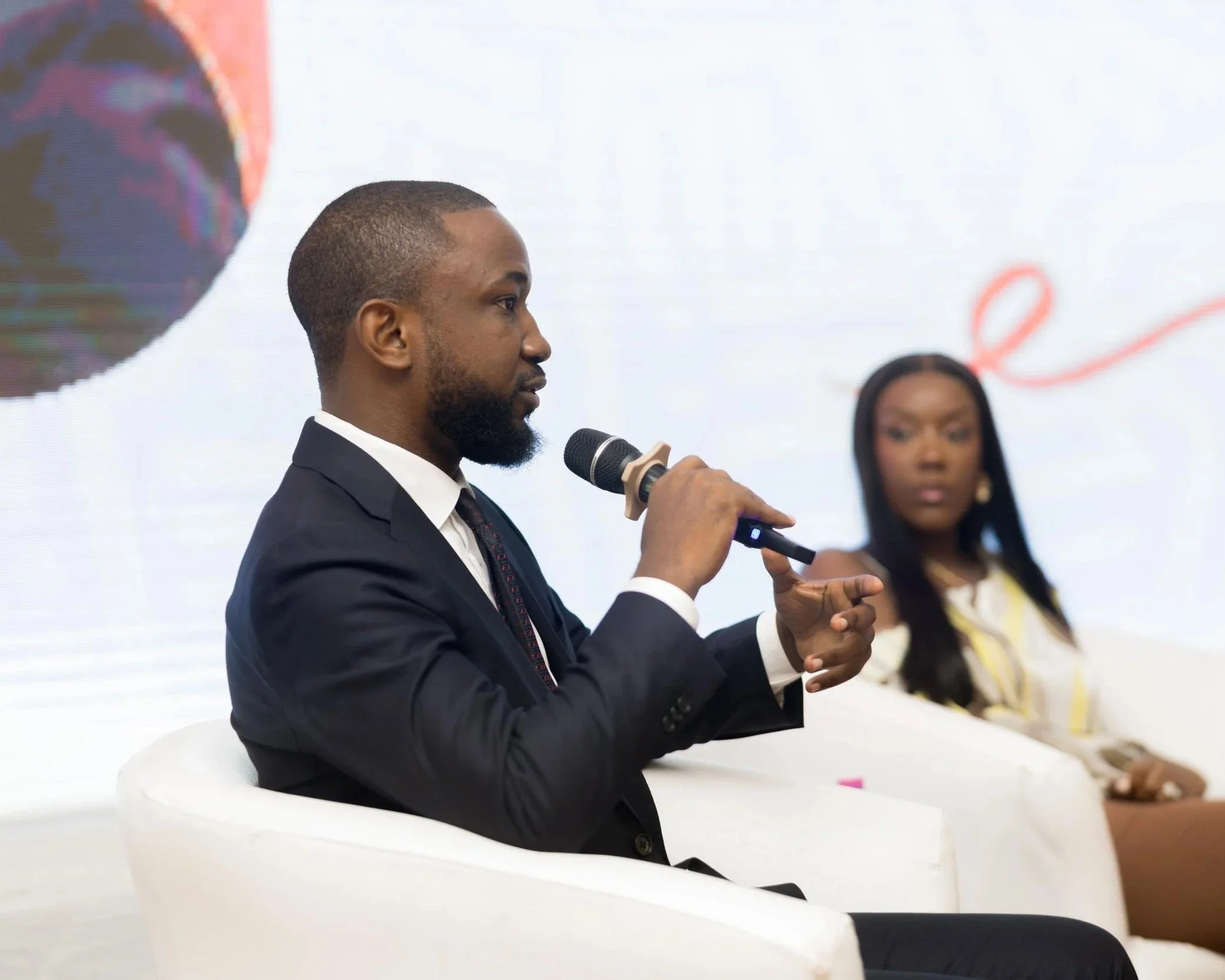 A man in a black suit and white shirt holding a microphone and speaking at an event. A woman in a yellow and white outfit is sitting in the background.