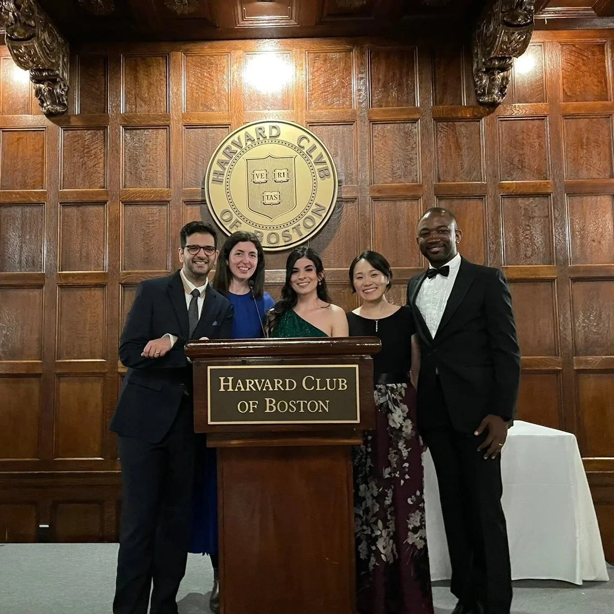 Group of five people dressed in formal attire standing behind a podium that reads 'Harvard Club of Boston' in a wood-paneled room with a Harvard emblem on the wall behind them.