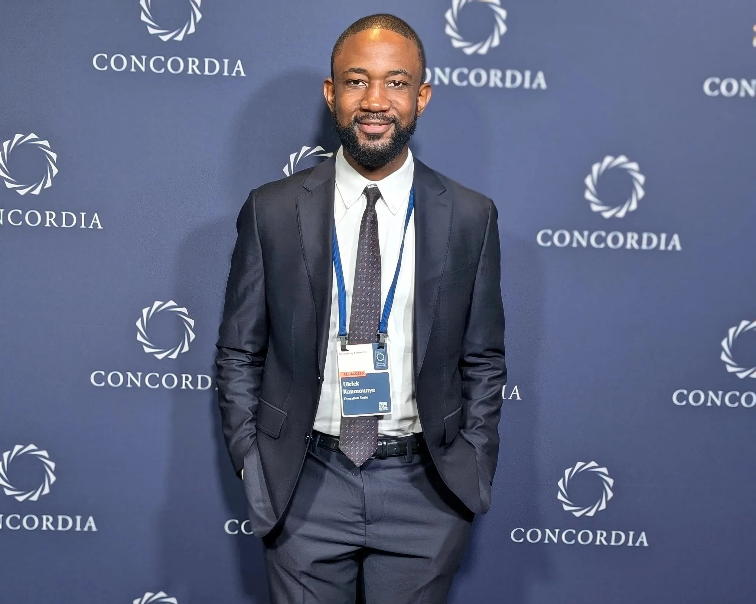 Man in a dark suit with hands in pockets standing in front of a navy blue backdrop with 'CONCORDIA' logos and text, wearing a white shirt, black tie, and an event badge.