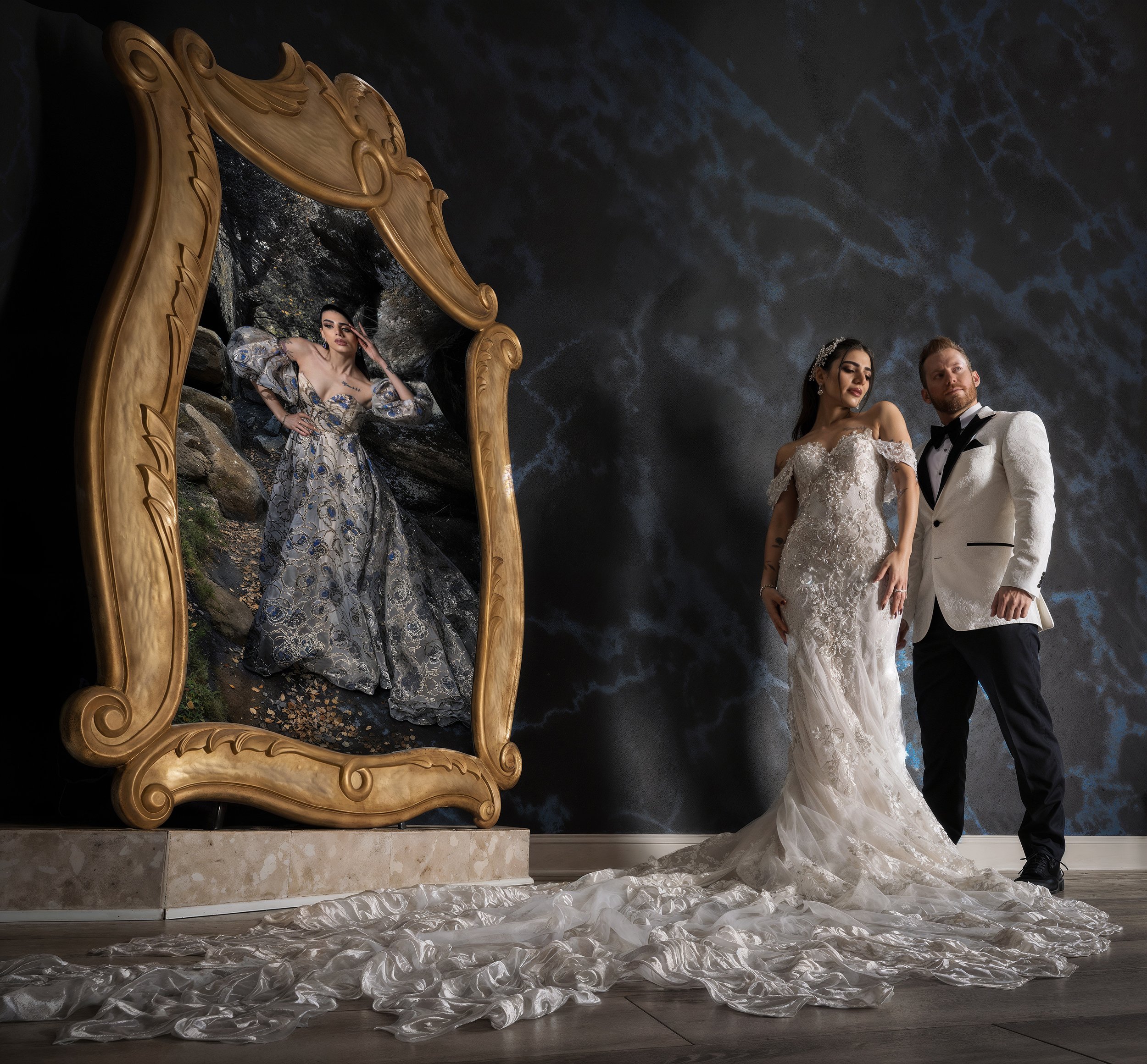 A bride and groom in wedding attire stand next to a large ornate mirror. The bride wears a white, off-the-shoulder lace wedding gown with a long train, and the groom wears a white tuxedo jacket with black pants. They pose confidently with a dark, sto