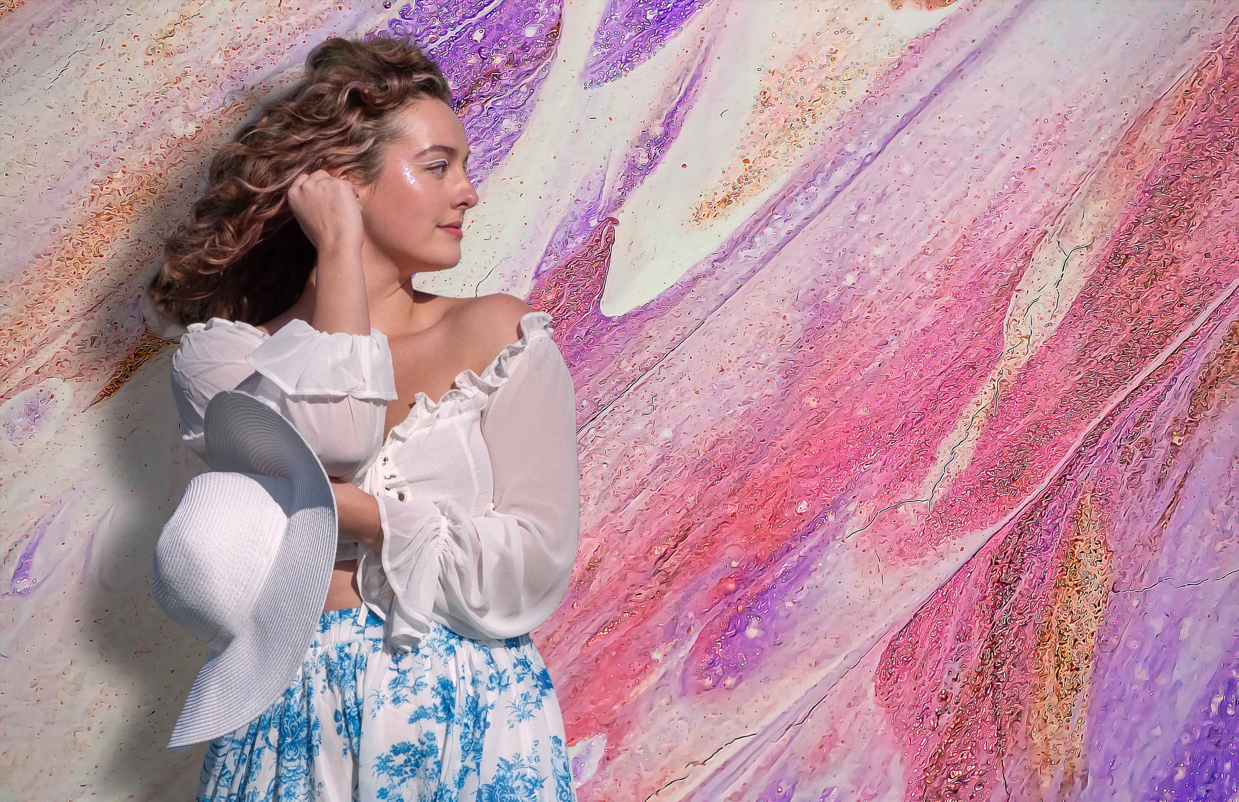 A woman with curly hair and a white off-shoulder blouse is lying on a colorful abstract background, holding a white hat and looking to her left.