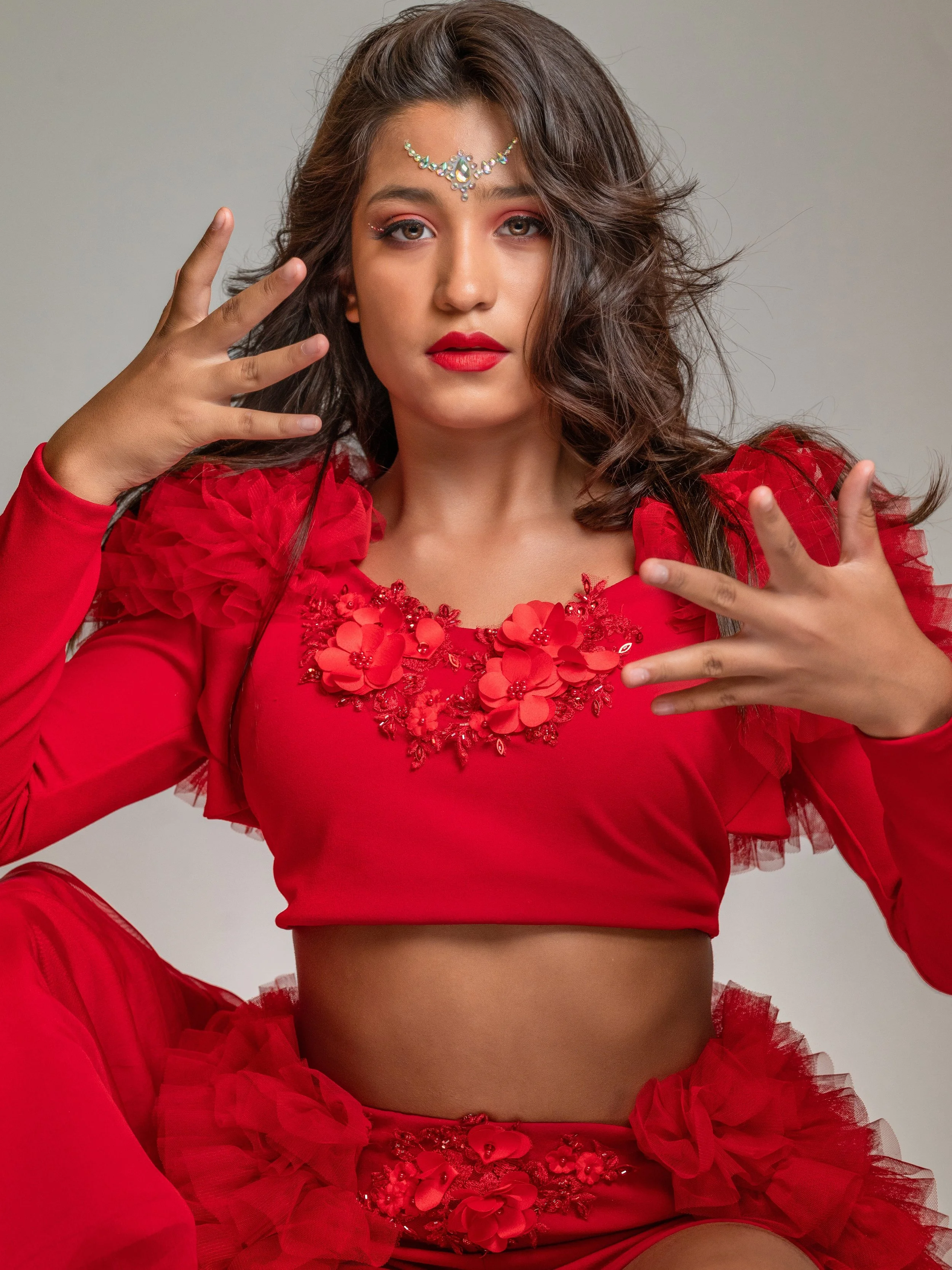 A woman in a red dress with floral embroidery on the chest, posing with her fingers near her face and wearing a decorative headpiece with jewels. She has long, wavy brown hair and red lipstick.