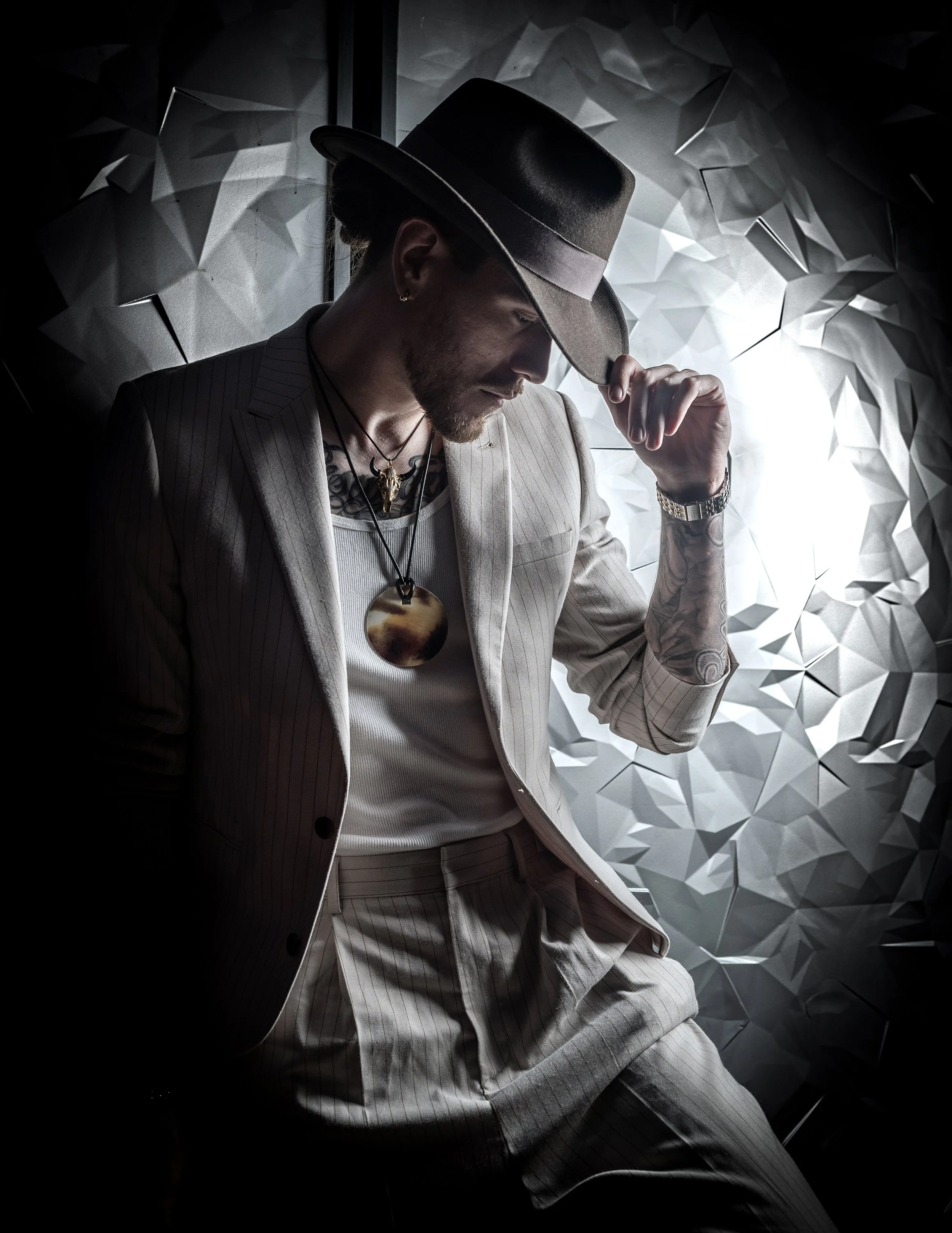 A man dressed in a beige pinstripe suit and matching shorts, wearing a black hat, silver watch, and layered necklaces, is standing against a textured geometric wall, with his head slightly bowed and hand adjusting his hat.