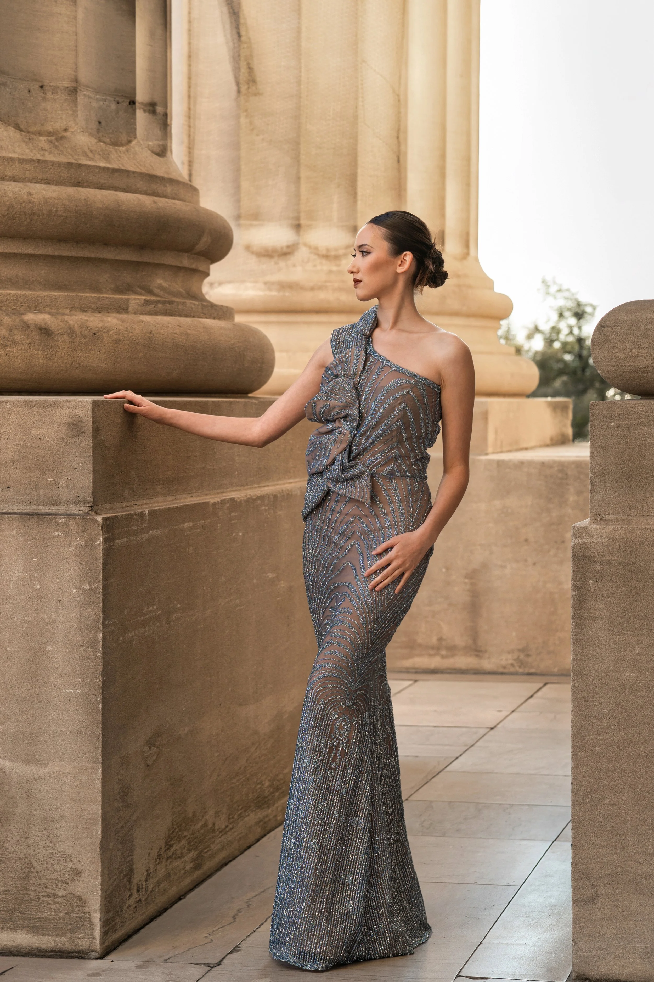 A woman in a shimmering, one-shoulder silver gown standing beside large stone columns at a historic site.