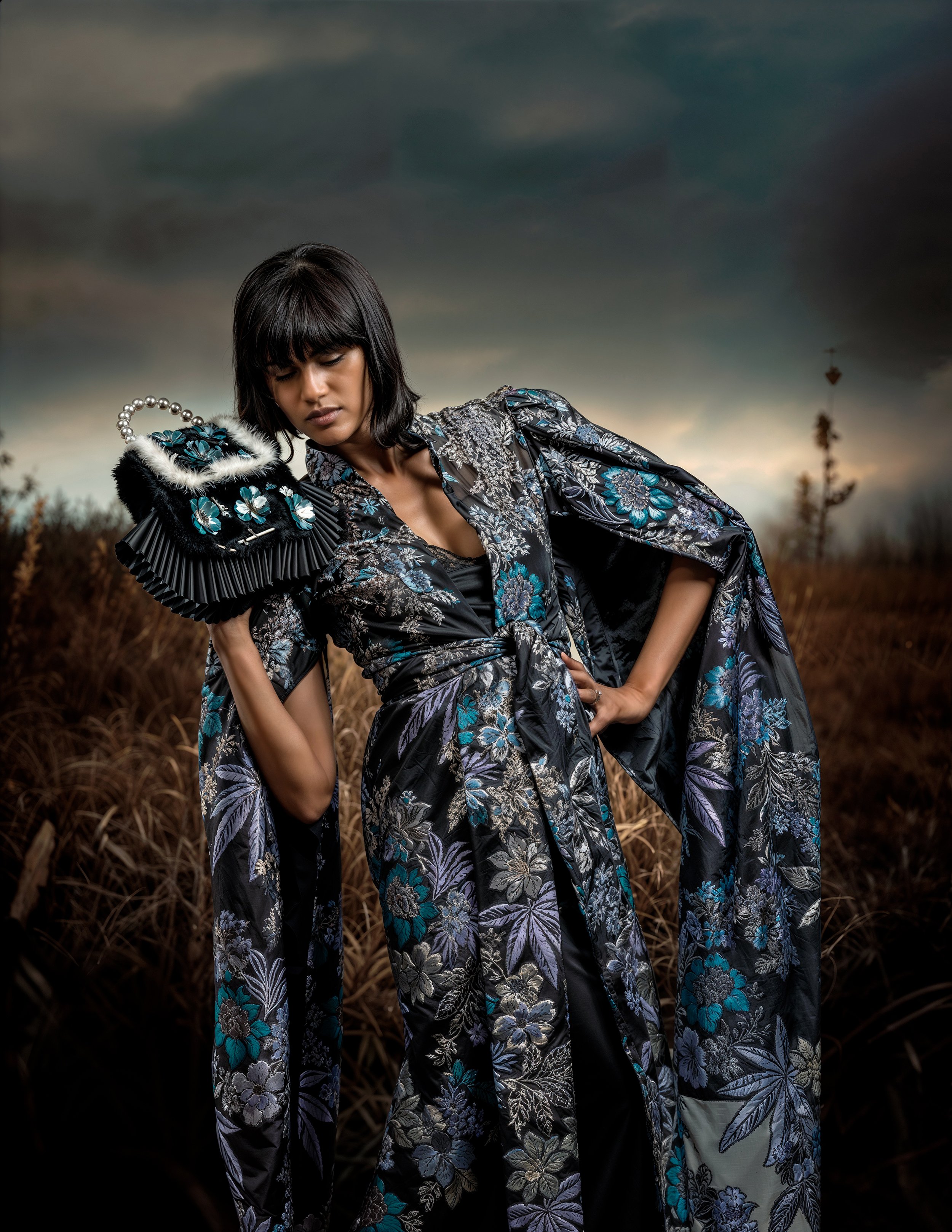 A woman in a floral kimono-style outfit standing in a field of tall, dry grass under a dark, cloudy sky, with a posed, contemplative expression.