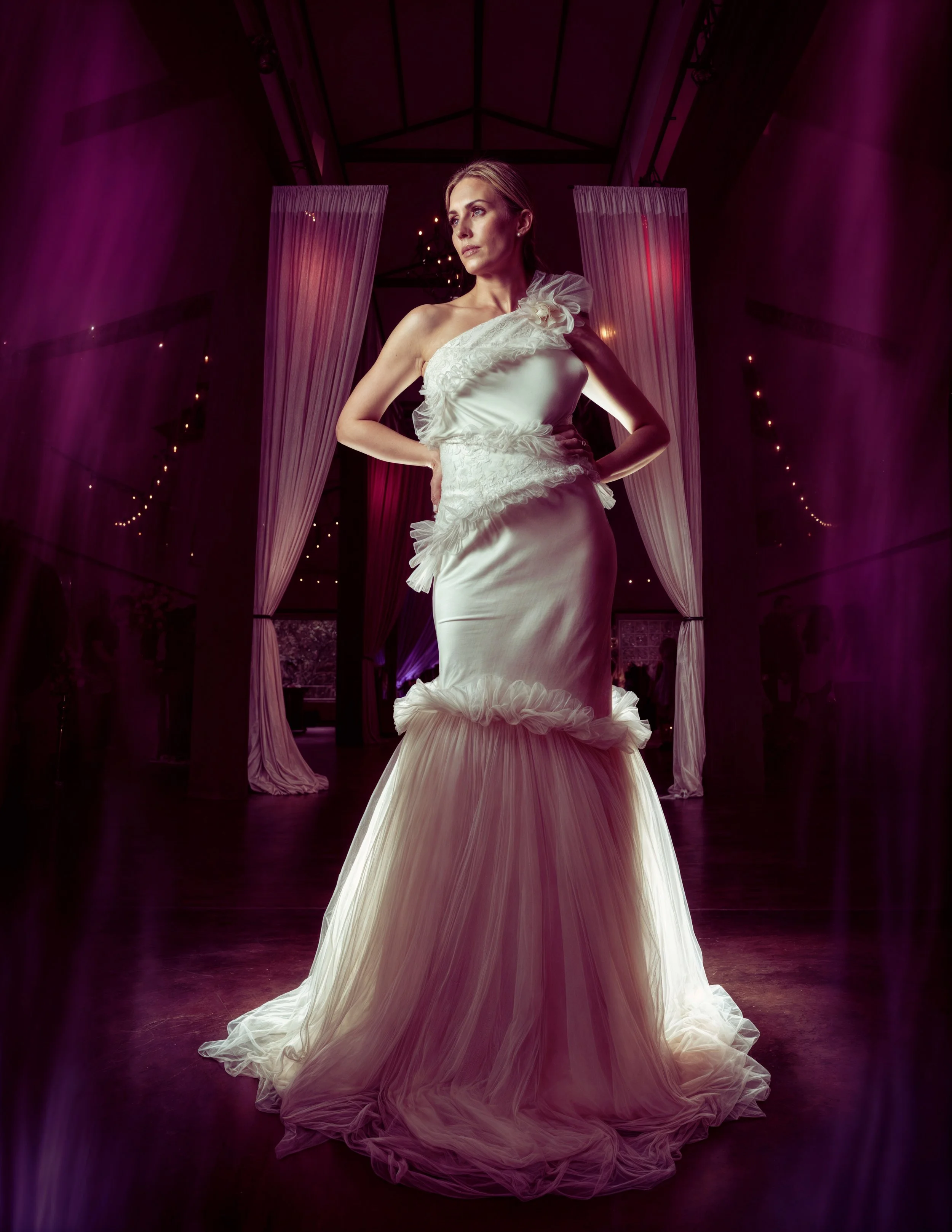 A woman in a white, ruffled, strapless wedding gown with a fitted bodice and a long flowing train, standing in a dimly lit room with pink and purple lighting, drapery, and string lights in the background.