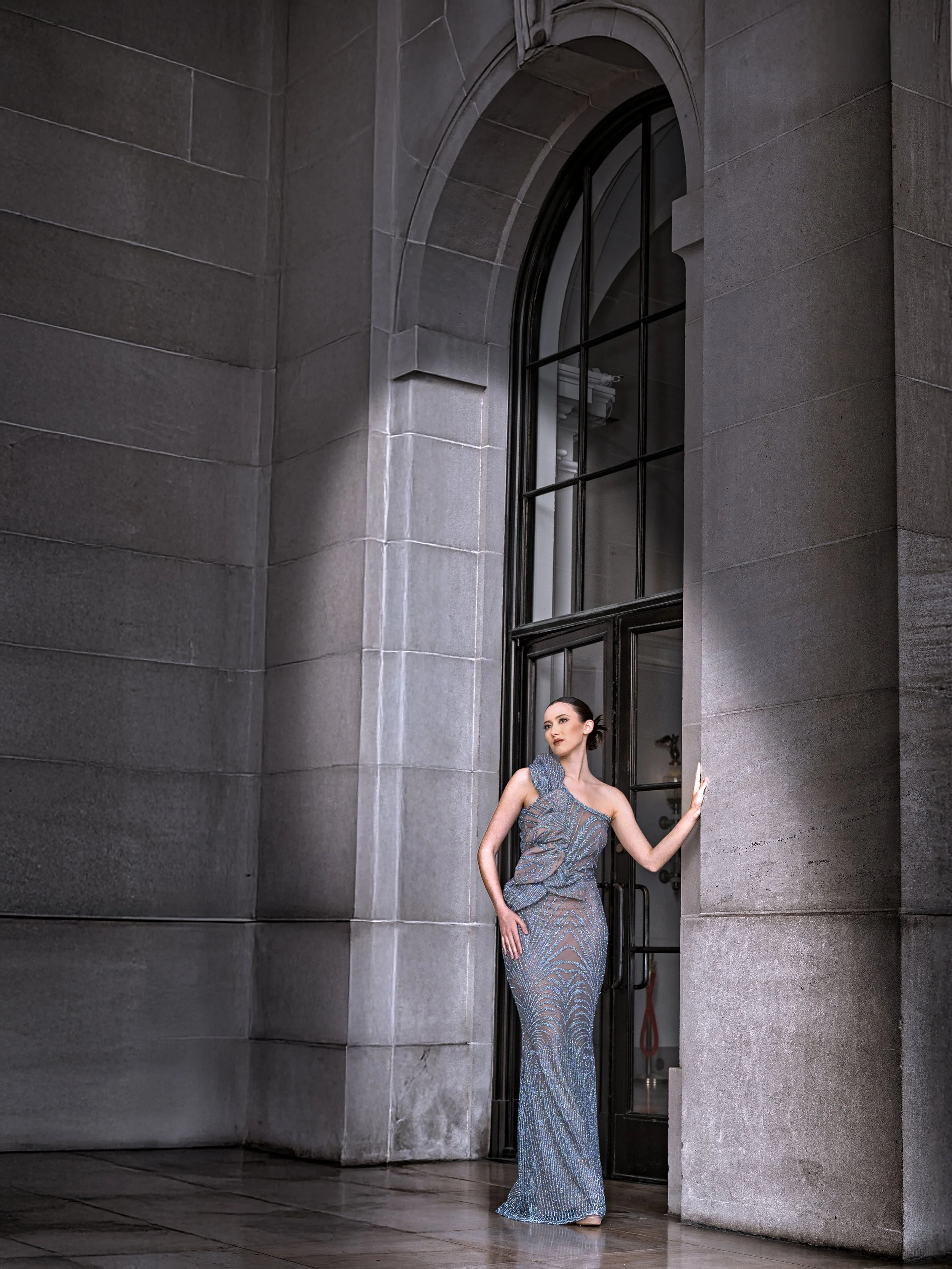 A woman in a form-fitting, metallic blue gown stands in an interior setting with large gray stone walls and a tall arched window, touching the wall with her right hand and looking to her left.