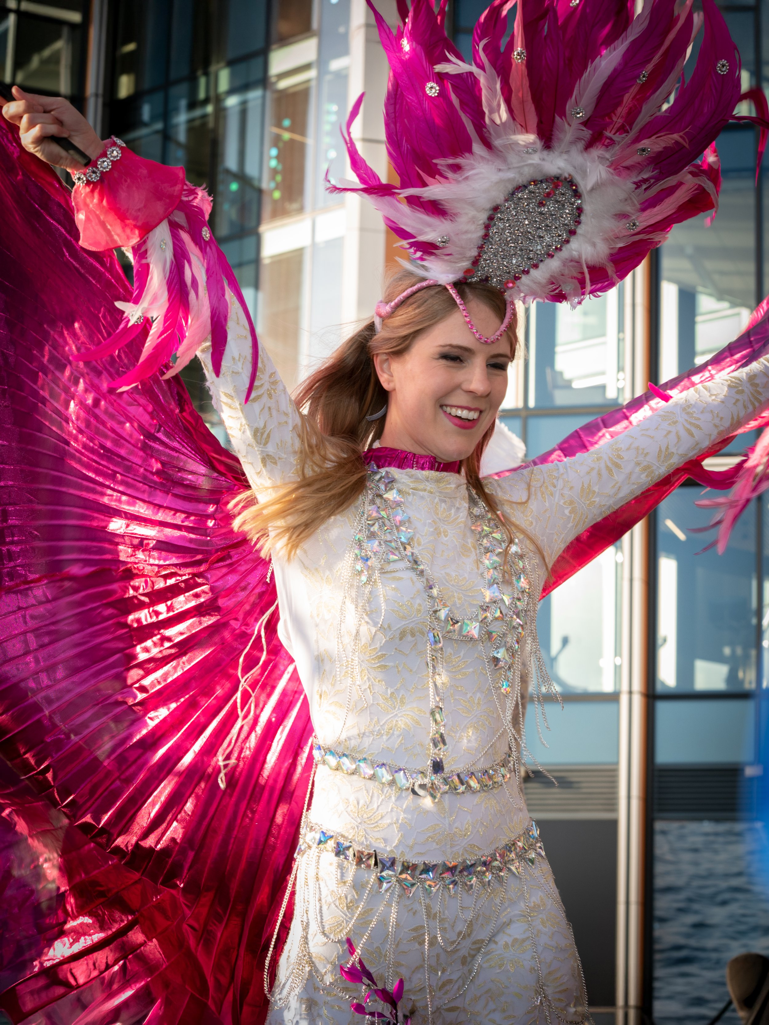 A woman dressed in a white and gold outfit with silver embellishments, wearing a large pink feathered headdress, waving her arms with a smile during a celebration or parade.