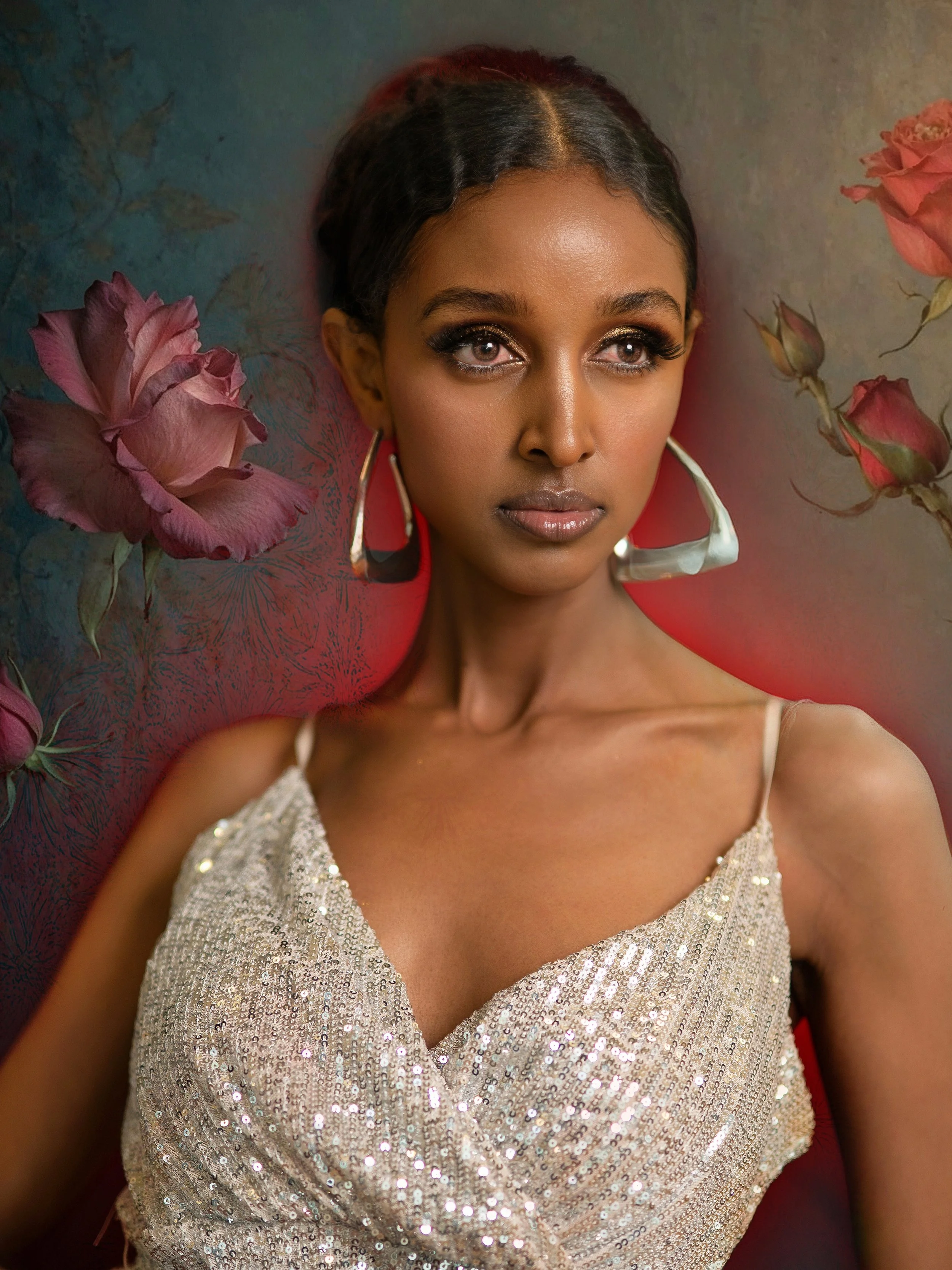 A woman with dark hair styled in waves, wearing a sparkly, sleeveless dress and large hoop earrings, posed against a colorful background with pink roses and greenery.