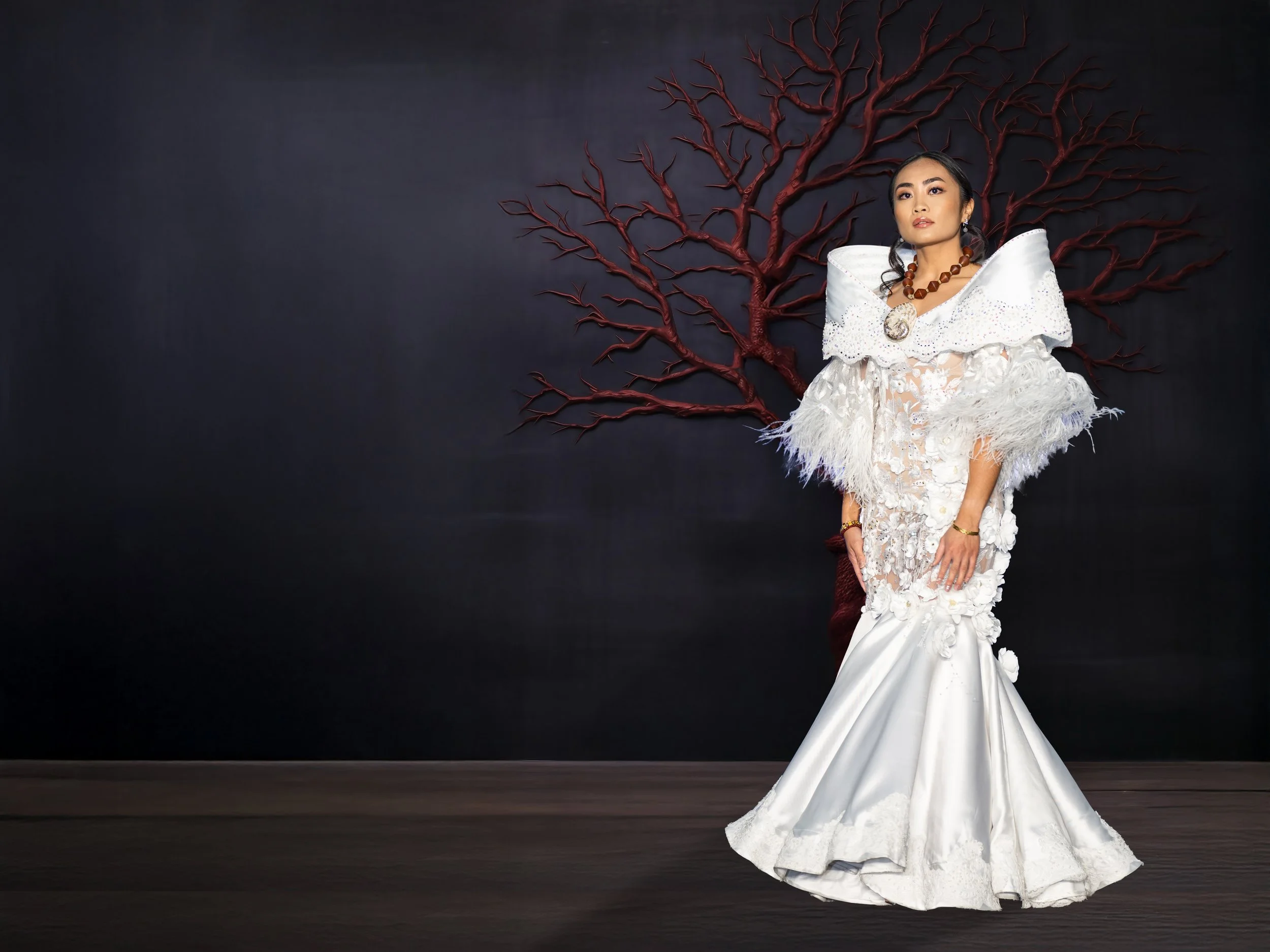 A woman in a white, elaborate dress with textured details and feathered sleeves posing in front of a dark wall with a red painted tree in the background.