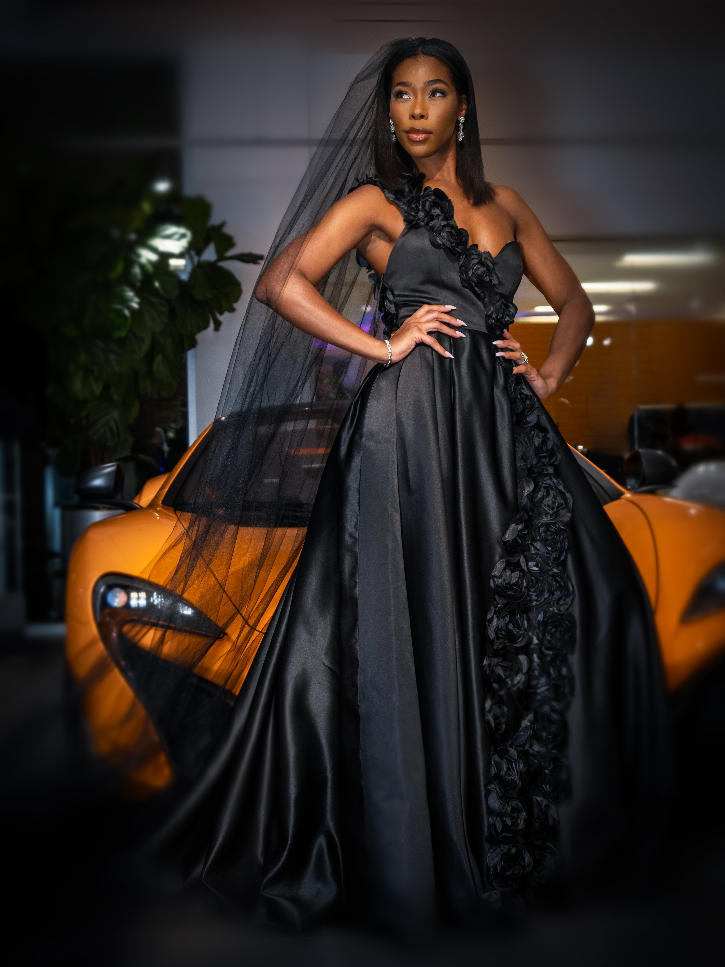 A woman in a black gown with floral details, standing next to a yellow sports car inside a modern building.