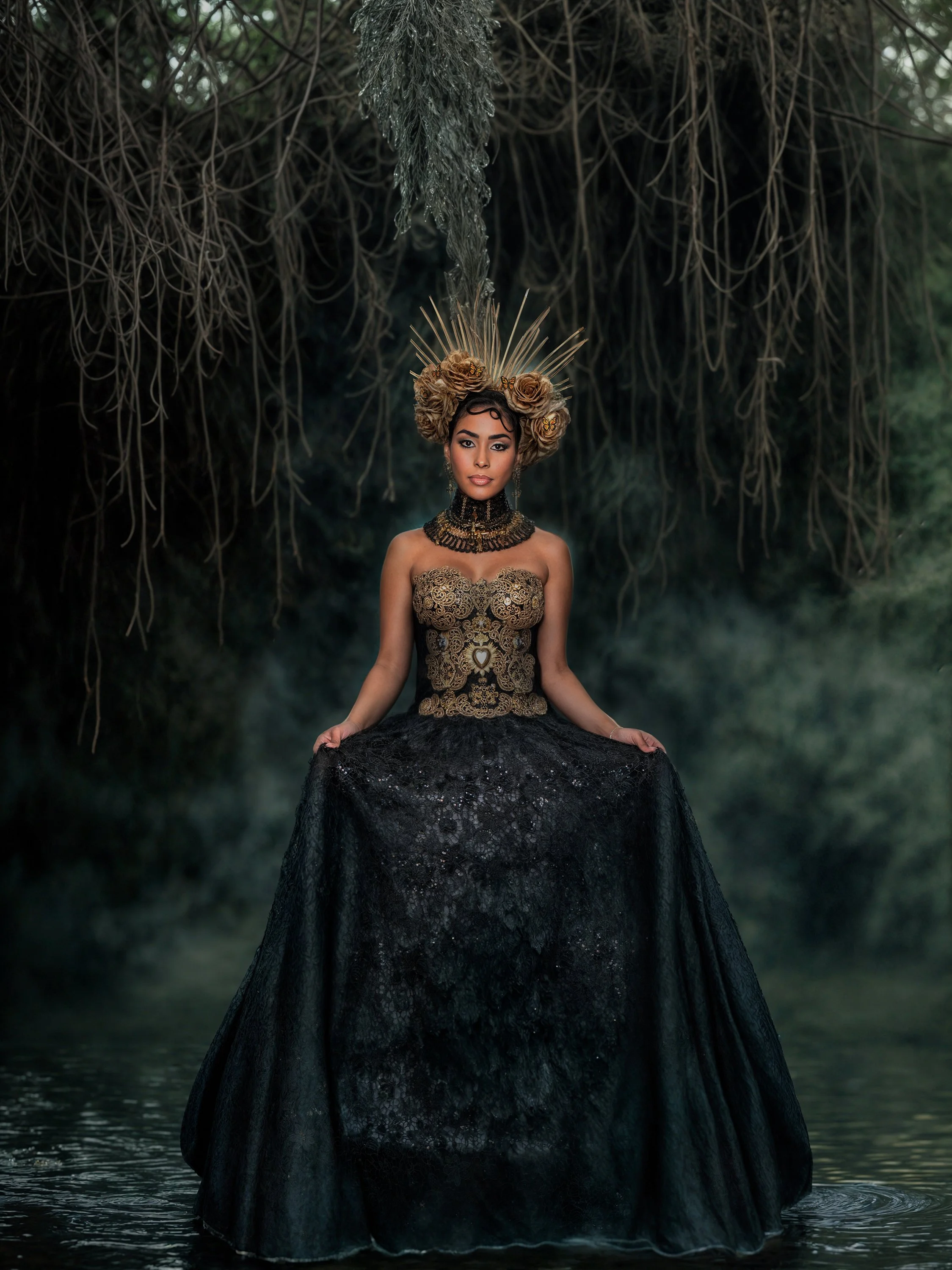 A woman dressed in an elaborate black and gold gown standing in water beneath overhanging tree roots, with a decorative headdress and jewelry.