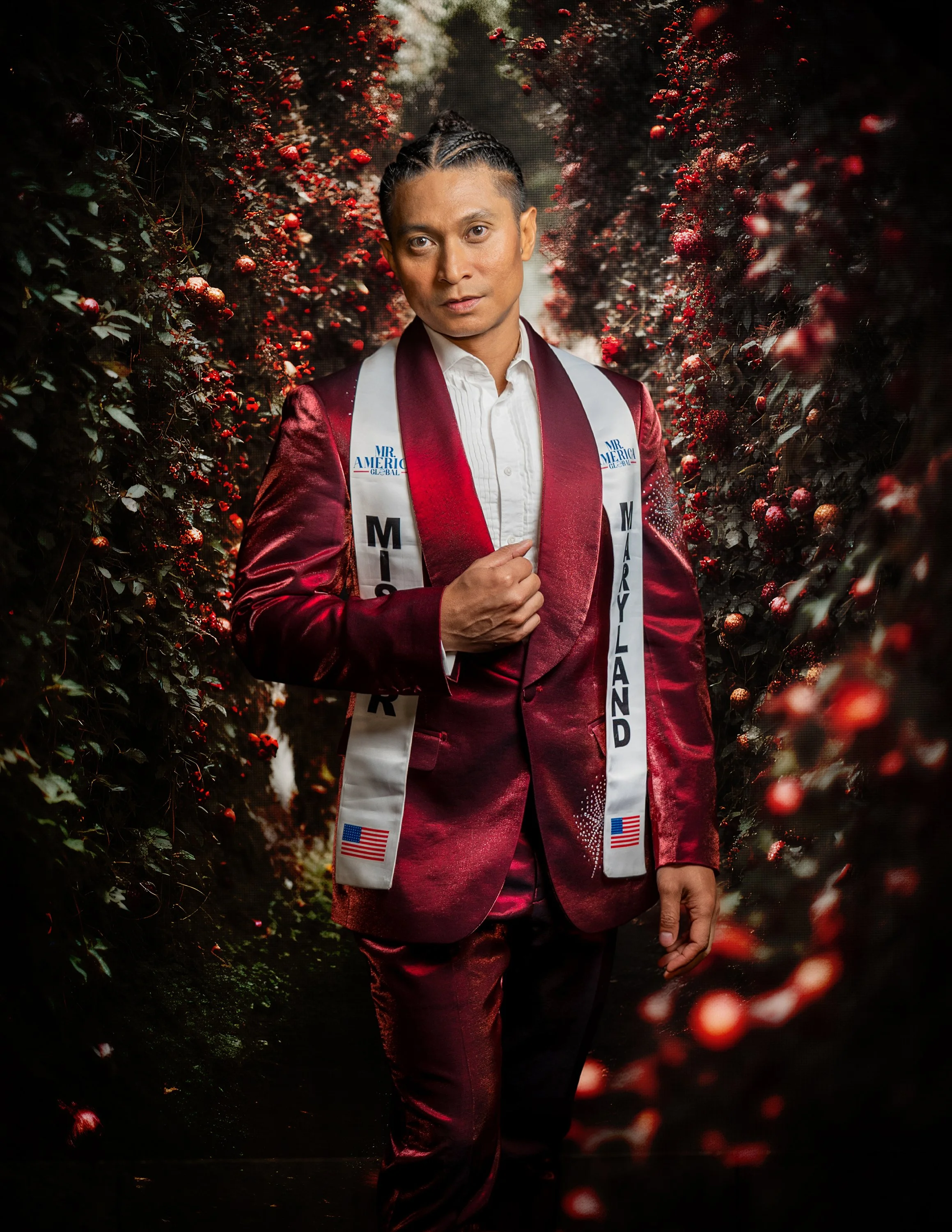 A man dressed in a shiny red suit and white shirt standing in between rows of red berry bushes. He is wearing a sash that says 'Mr. America' and 'Maryland' along with American flags.