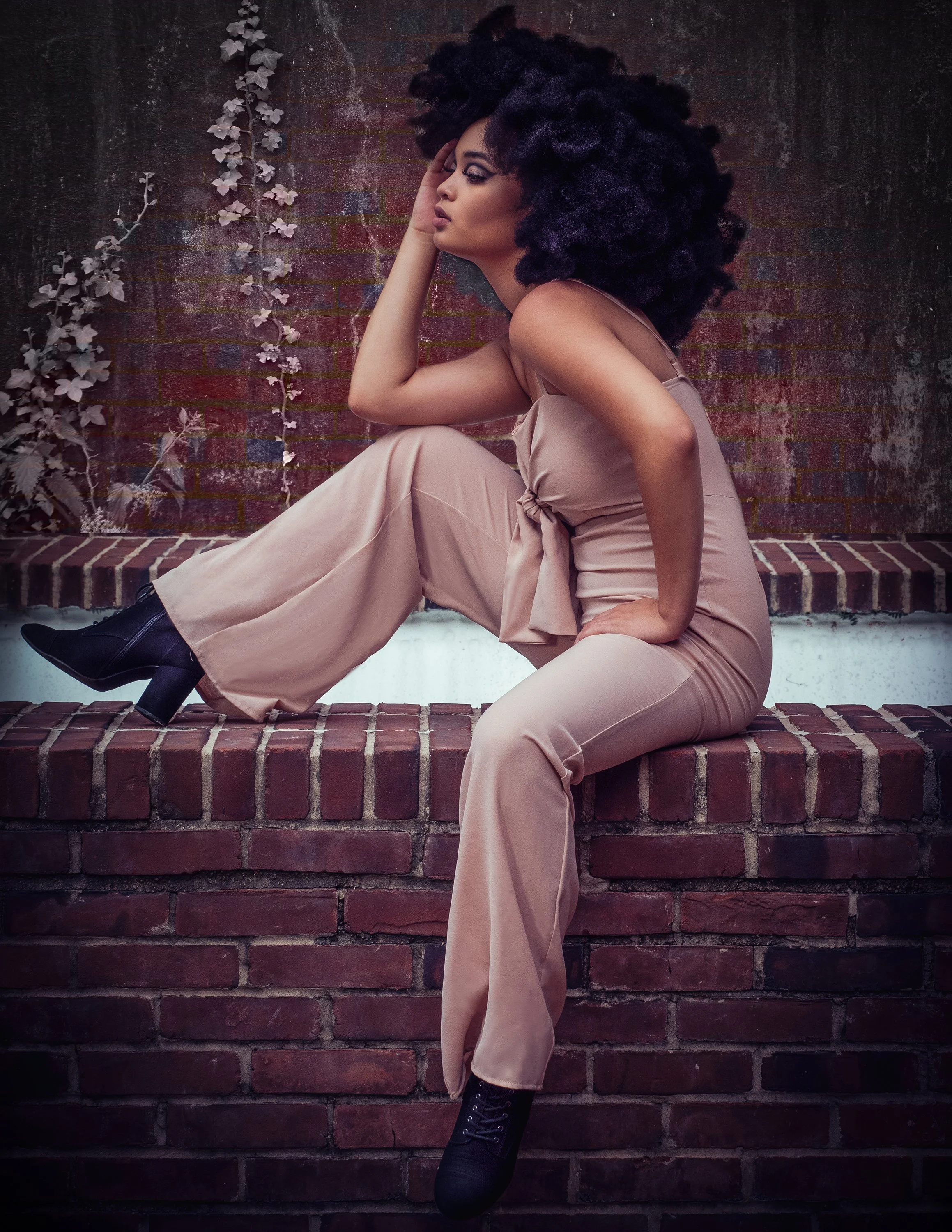 A woman with large curly hair sits on a brick ledge against a brick wall, wearing a beige jumpsuit and black ankle boots, with one hand resting on her face and the other on her hip.