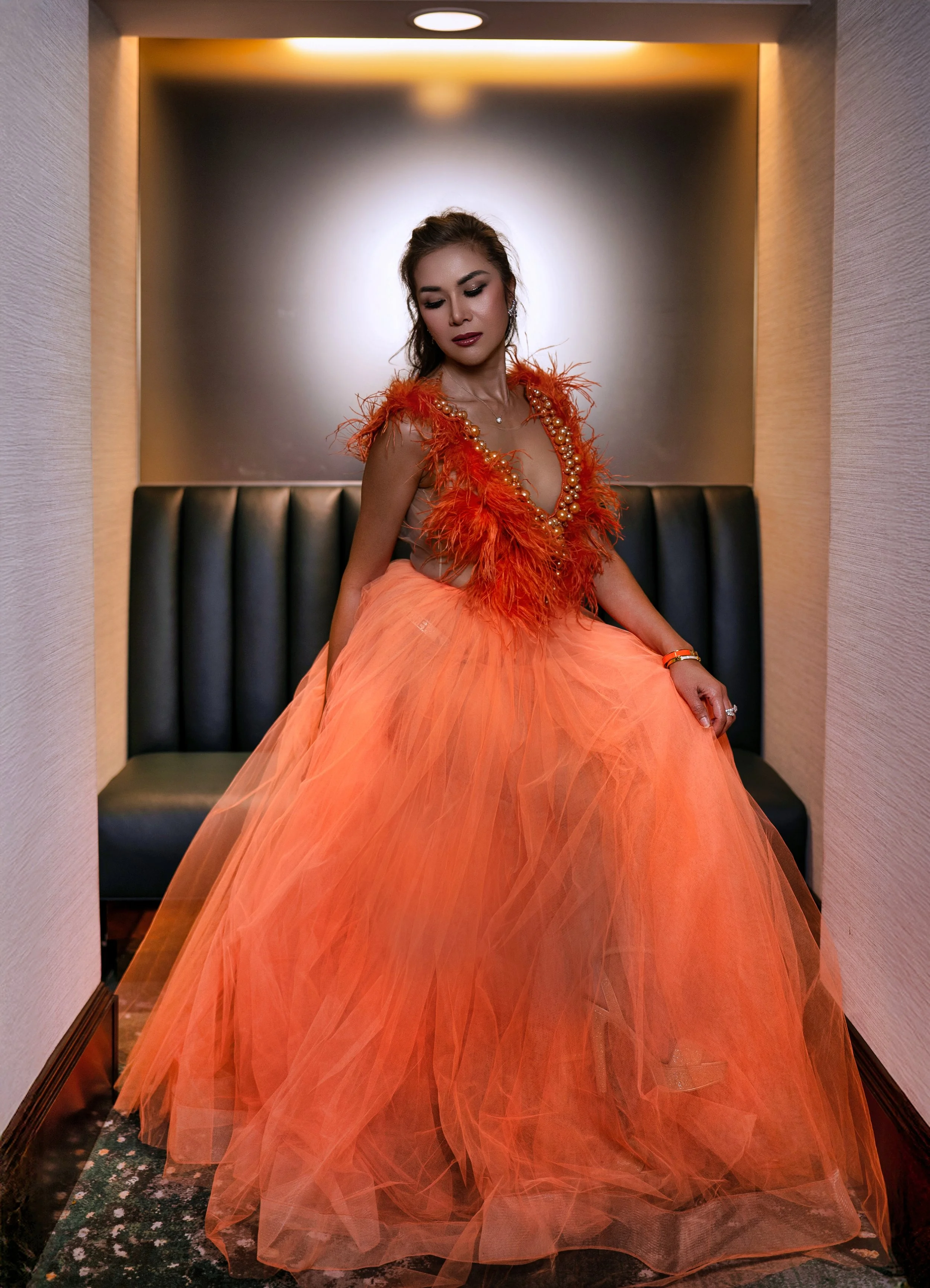 A woman wearing an orange gown with feathered embellishments and a tulle skirt sitting on a black bench in a small alcove with beige walls and a ceiling light.