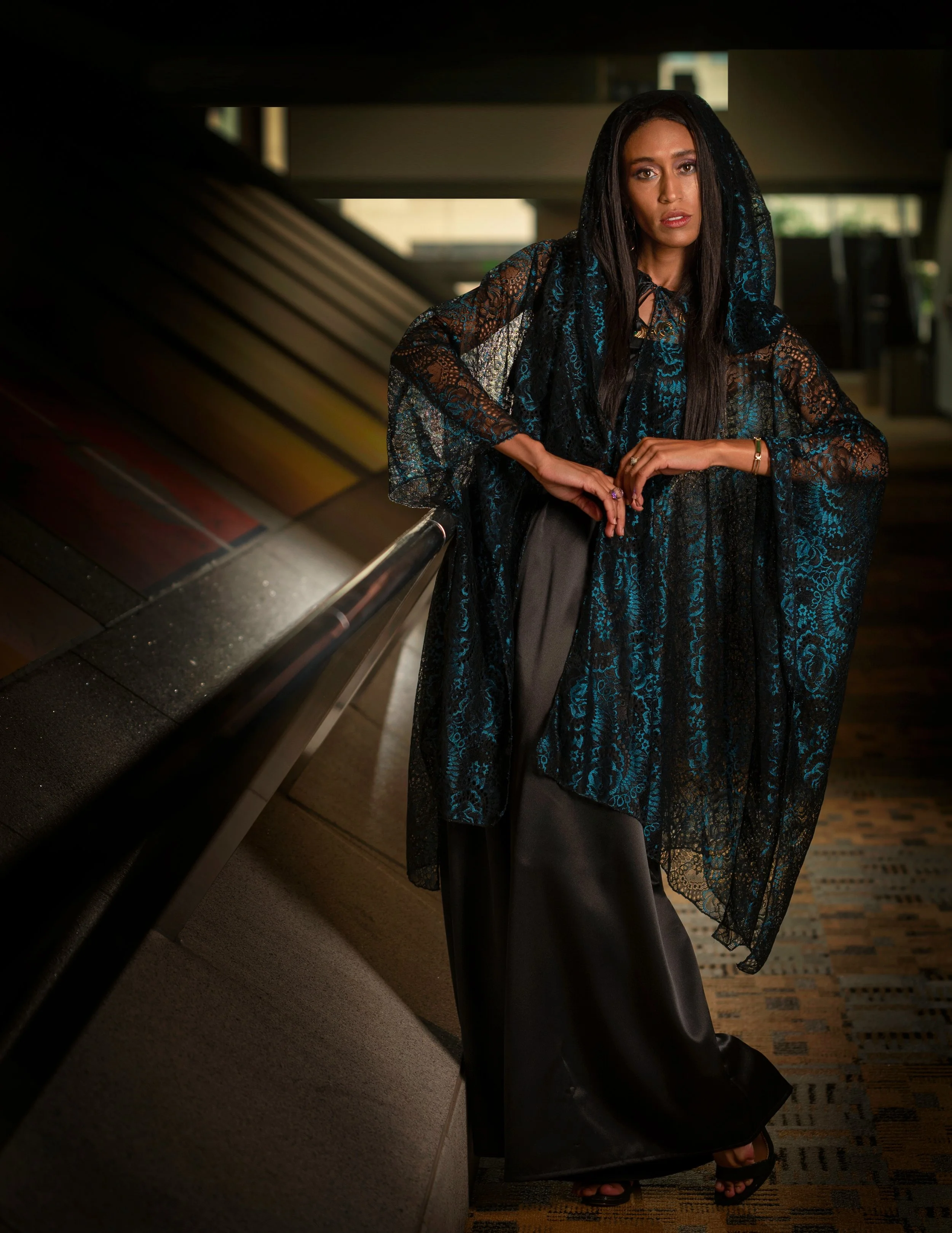 A woman with long dark hair wearing a black lace veil, a black lace top, wide-leg black satin pants, and high heels, standing inside a building near an escalator.