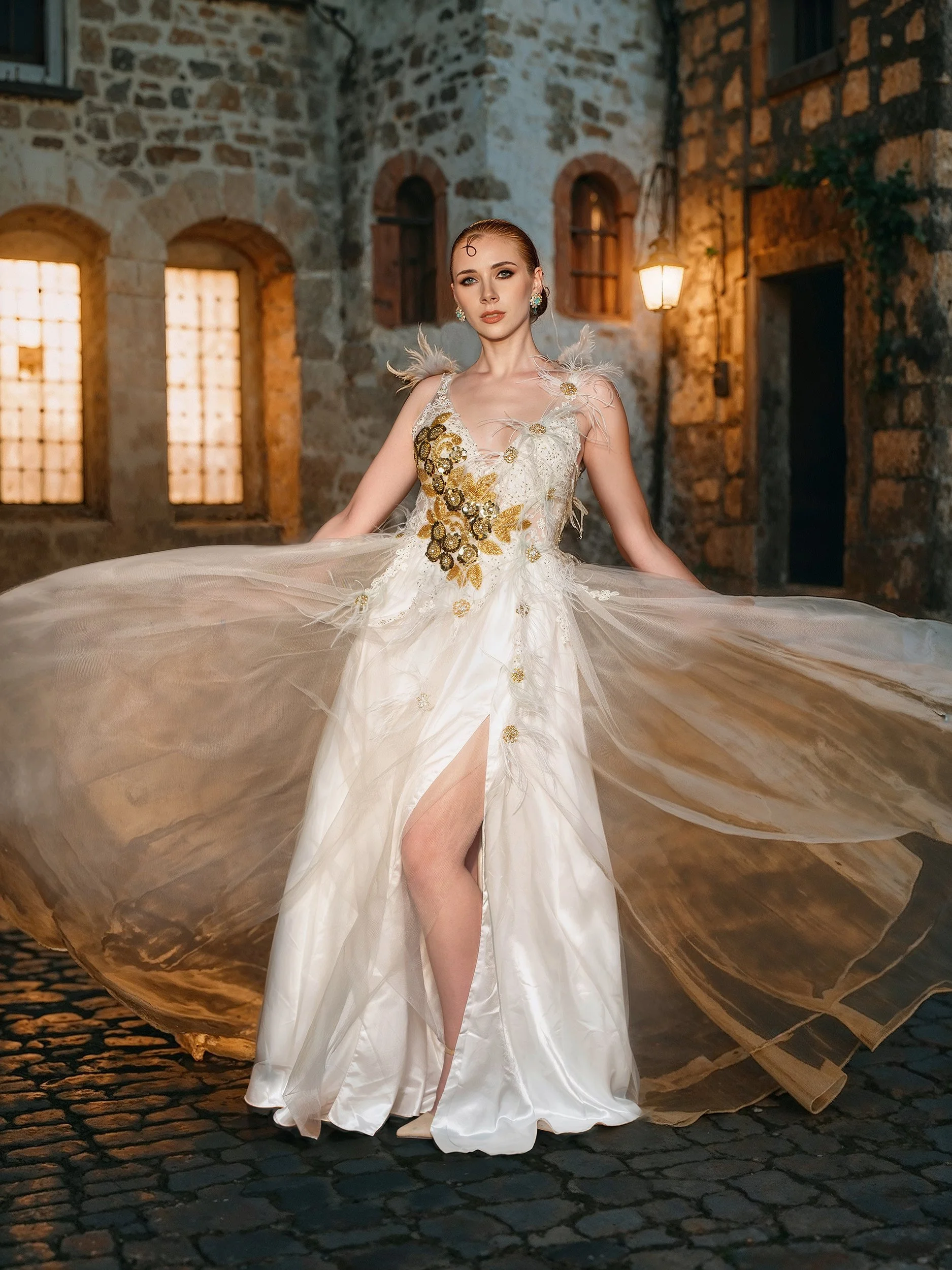 A woman in an elegant, gold-embellished white gown with a thigh-high slit, standing on a cobblestone street at dusk, with a stone building and warm streetlights in the background.