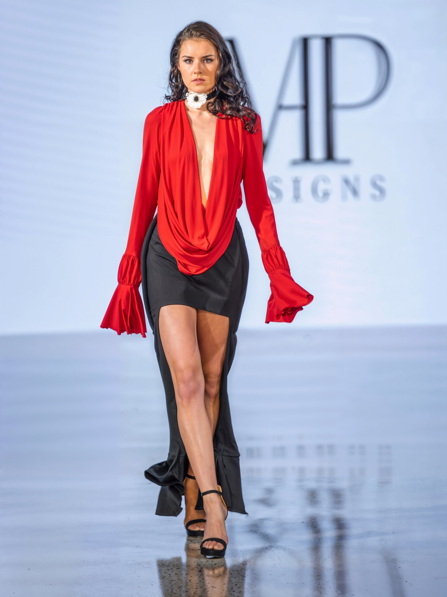 A female model wearing a red deep V-neck top with long, ruffled sleeves and a black high-slit skirt walking on a runway.