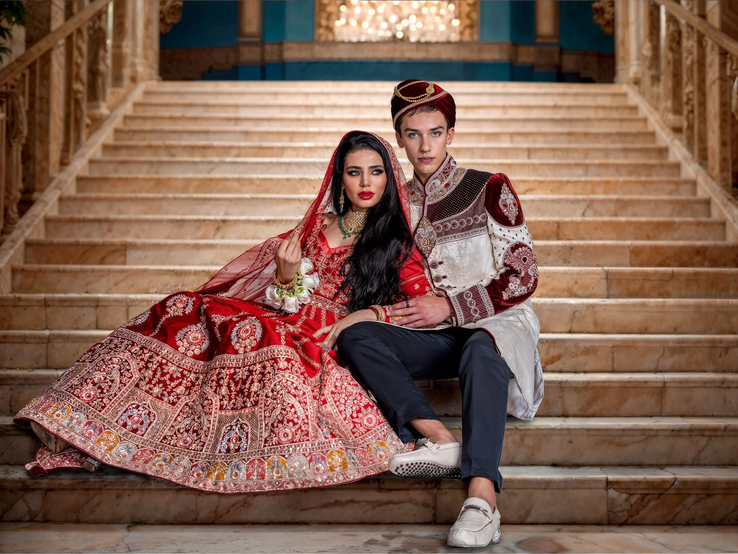 A couple dressed in traditional South Asian wedding attire seated on marble stairs in an ornate indoor setting.