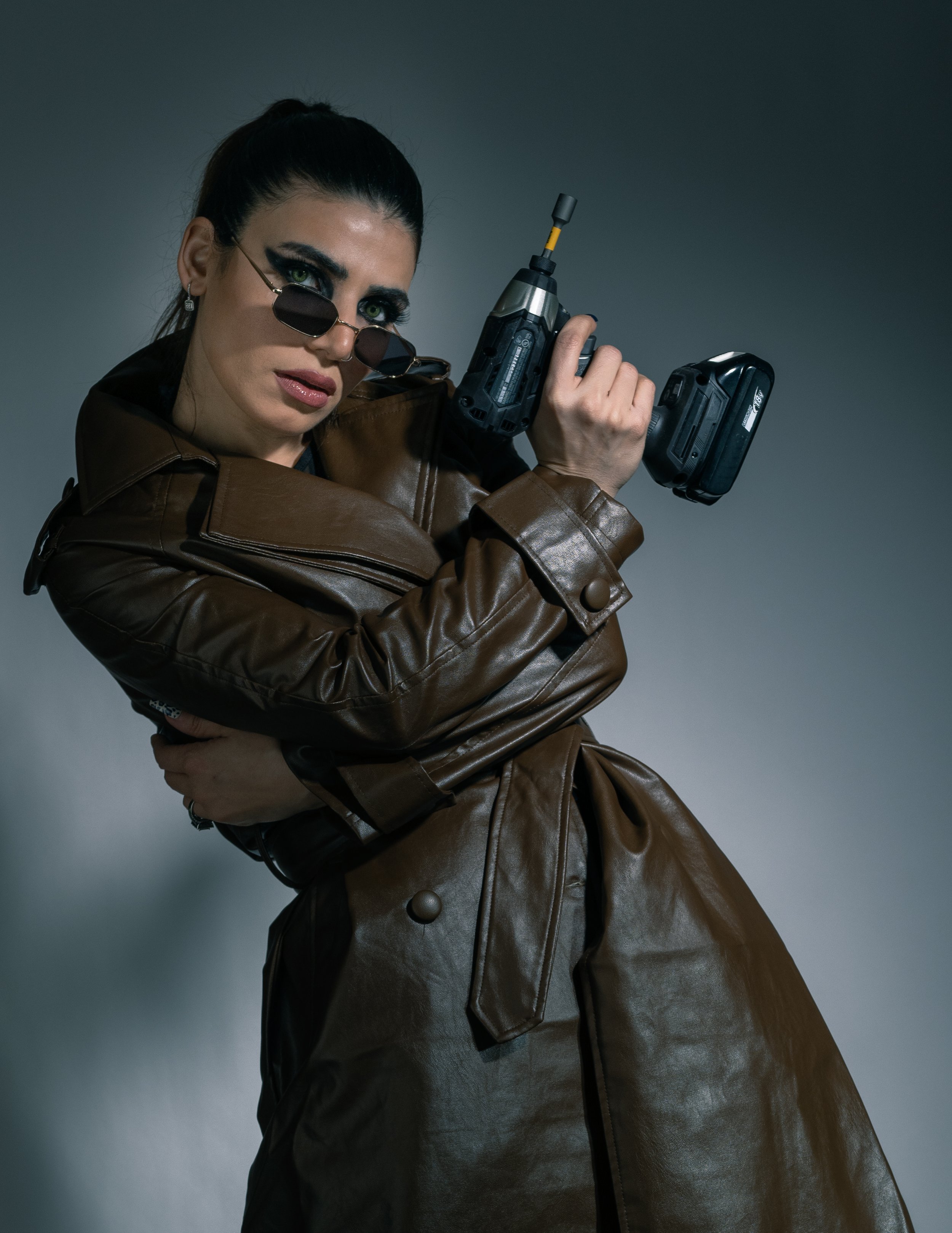 Woman with dark hair in ponytail, wearing sunglasses and a brown leather coat, holding a power drill with a camera attached.
