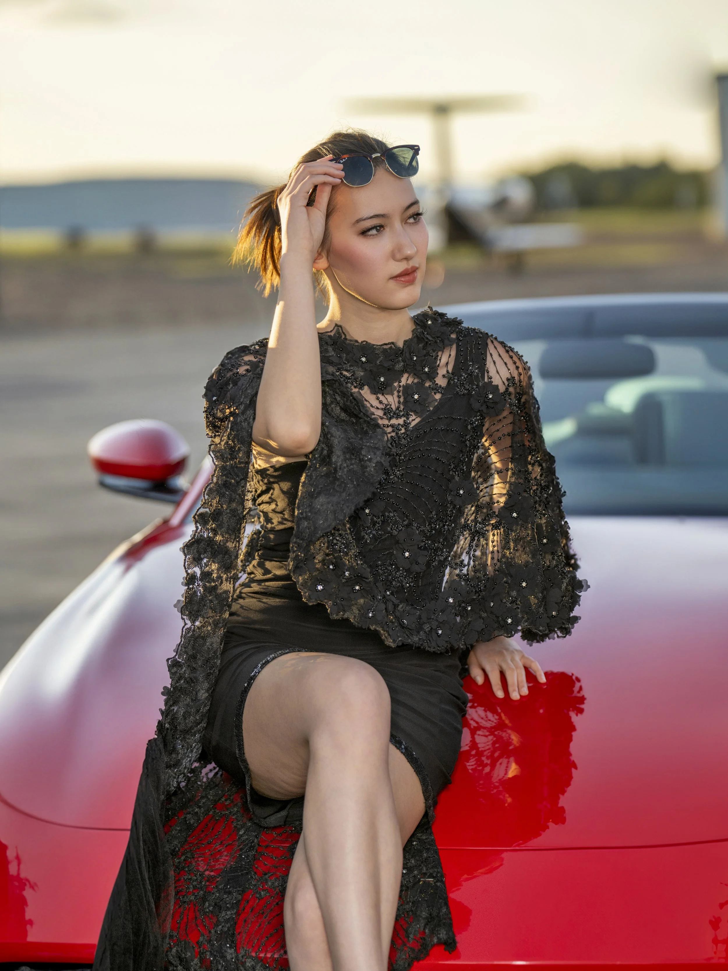 A young woman in a black dress with lace and embroidery details, sitting on the hood of a red convertible car, holding sunglasses in her hand, with short hair and a contemplative expression, during sunset.