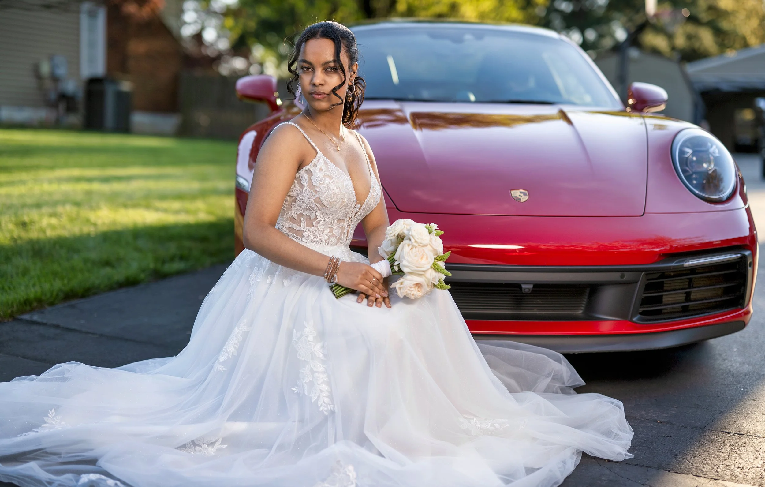 Bride in a wedding gown holding a bouquet of white roses, kneeling on a driveway in front of a red sports car.