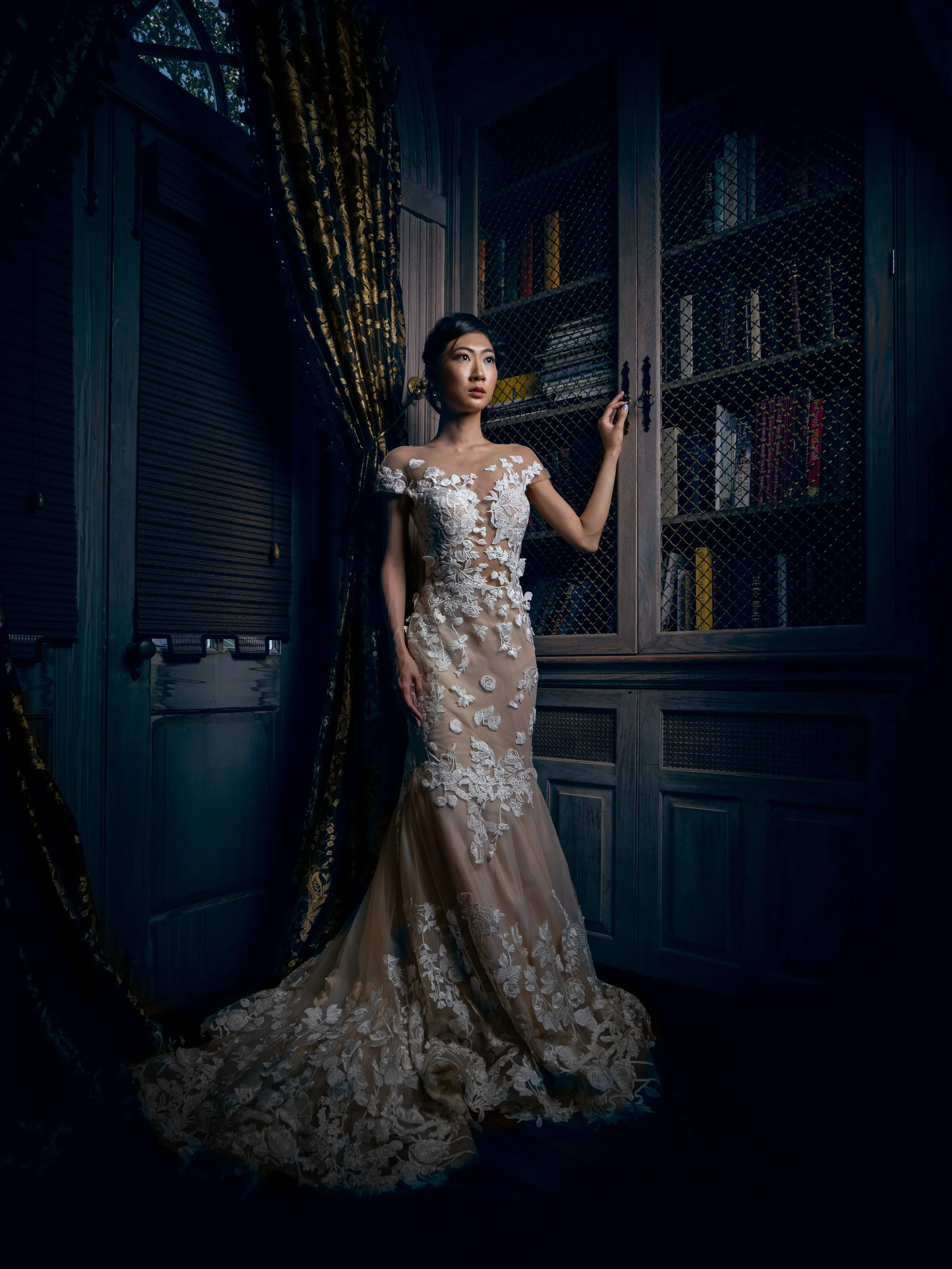 A woman in an elegant lace wedding dress standing in a richly decorated library with wooden bookshelves and dark curtains.