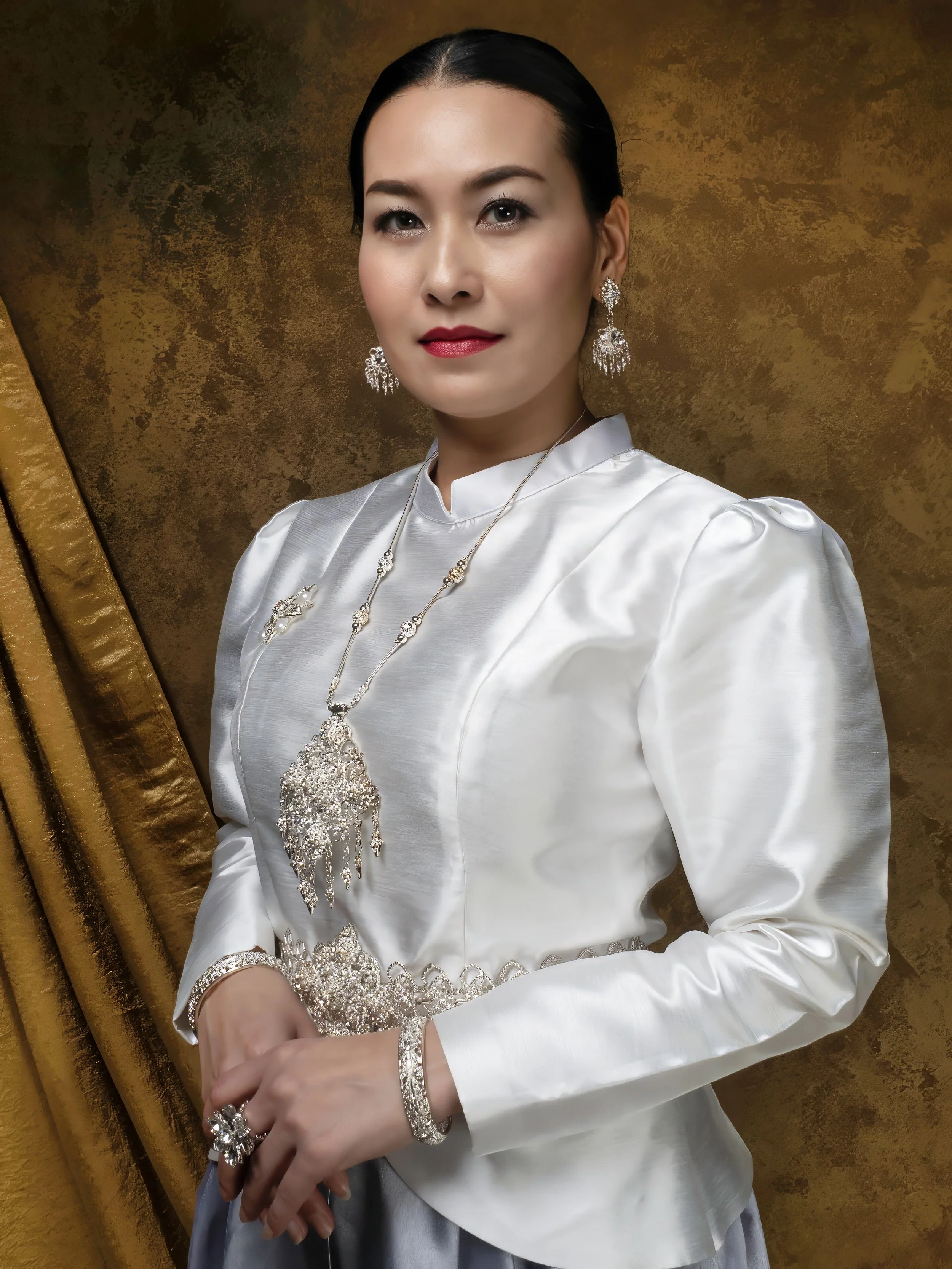 Portrait of a woman wearing a white satin blouse and silver jewelry, including earrings, a necklace, a bracelet, and a ring, standing against a gold and brown textured background.