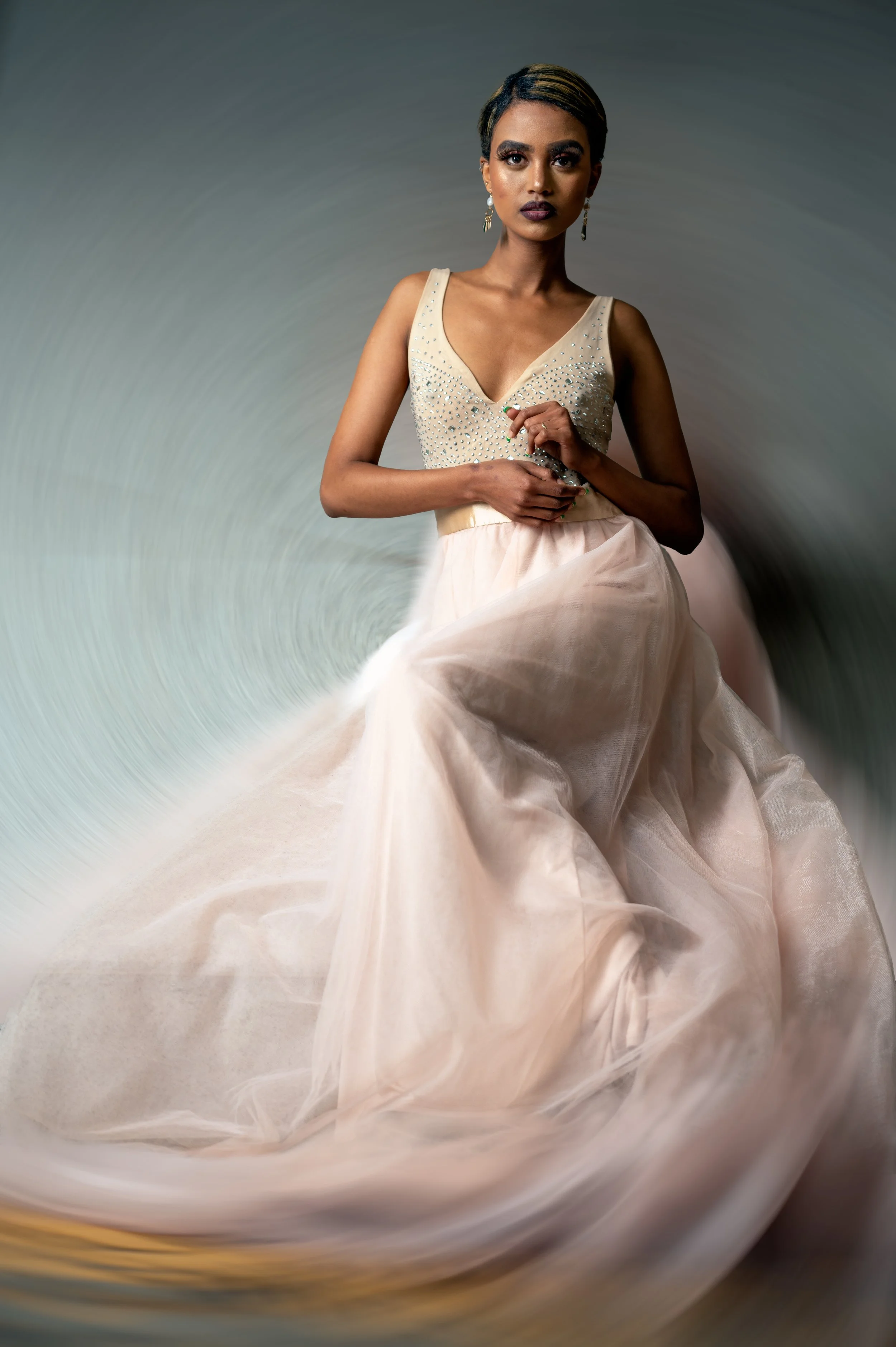 A woman wearing an elegant beige dress with rhinestones, sitting against a neutral background.