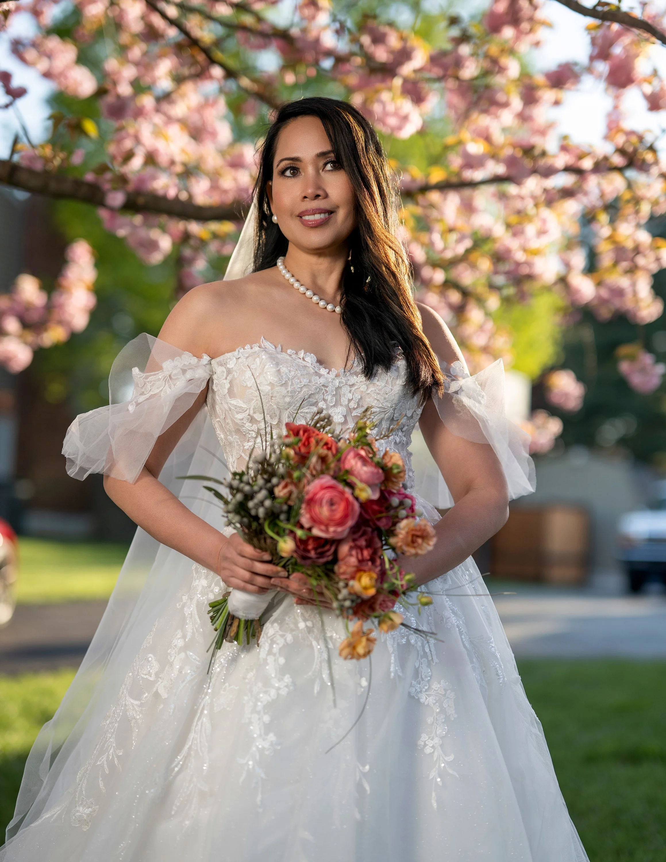 A woman in a wedding dress holding a bouquet of flowers outdoors during springtime, with pink cherry blossoms on the tree behind her.