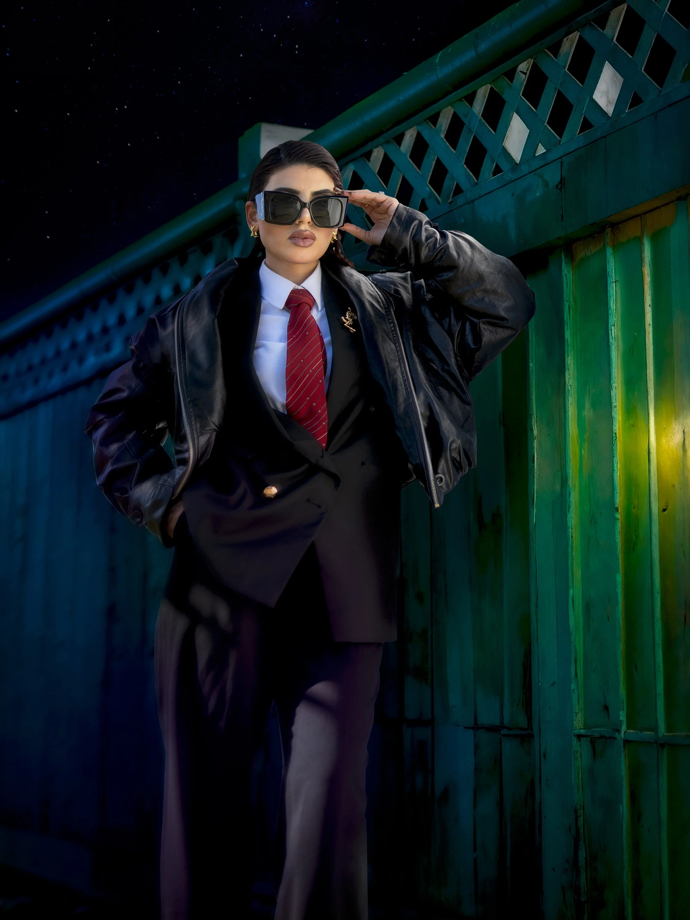 A stylish woman in a suit, leather jacket, and sunglasses standing against a railing at night.