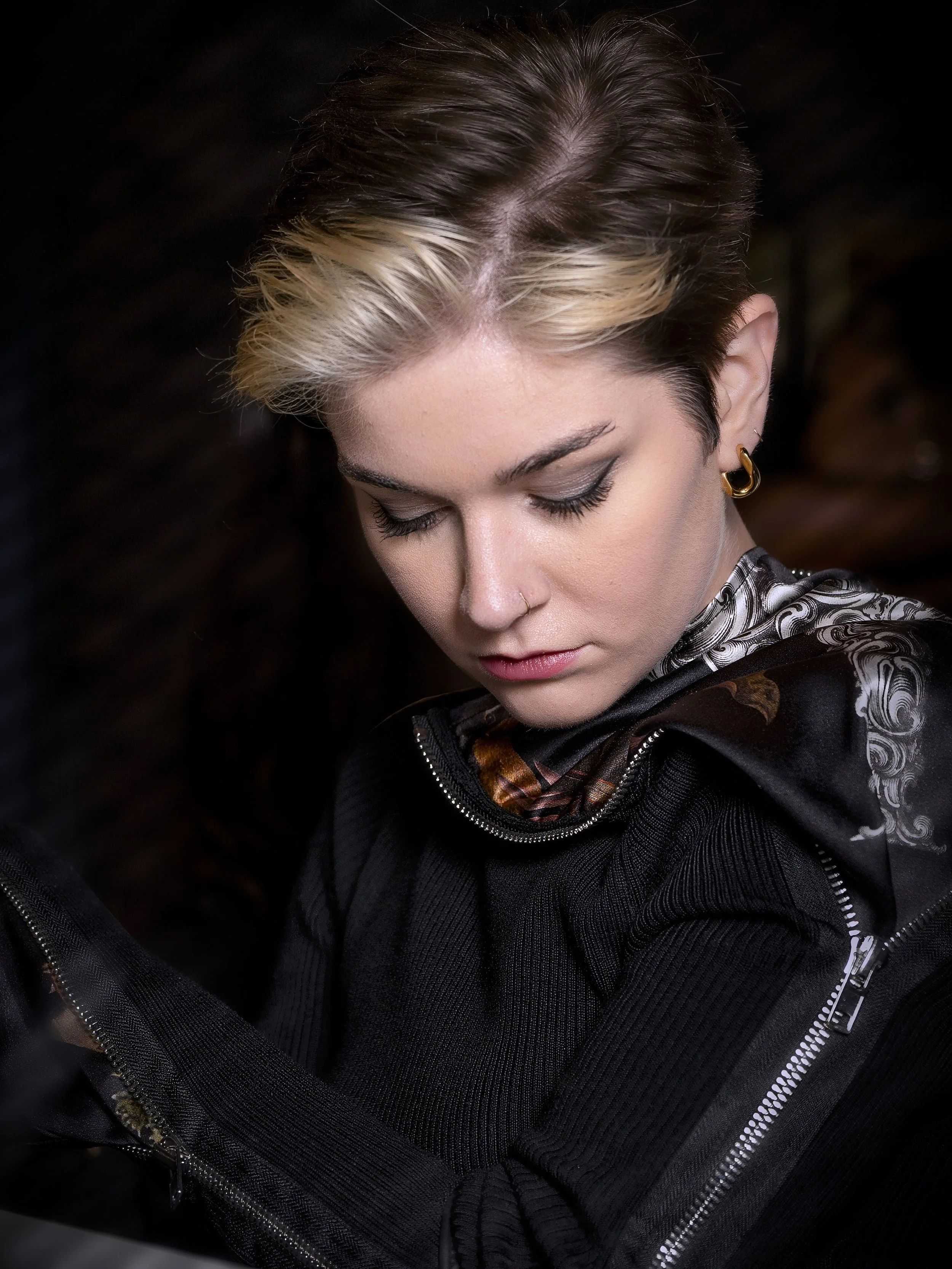 Young woman with short, dark hair and blonde streaks, wearing a black jacket with a patterned scarf, looking down with closed eyes and wearing gold hoop earrings.