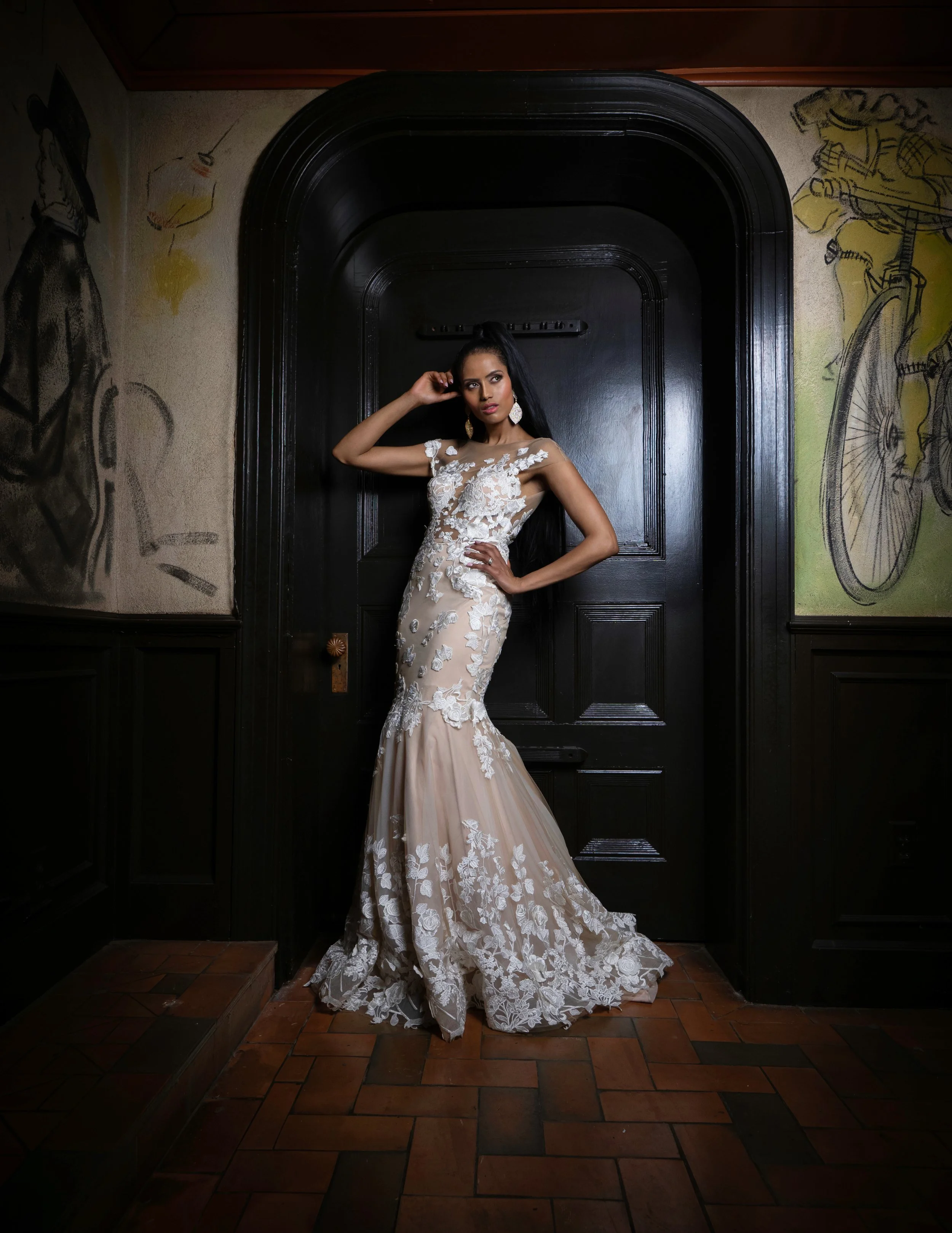 A woman in a beige and white lace wedding gown posing in front of a black door with mural art on its sides. The dress has floral embroidery and a fitted silhouette that flares at the bottom. She has long dark hair and is wearing large earrings, stand