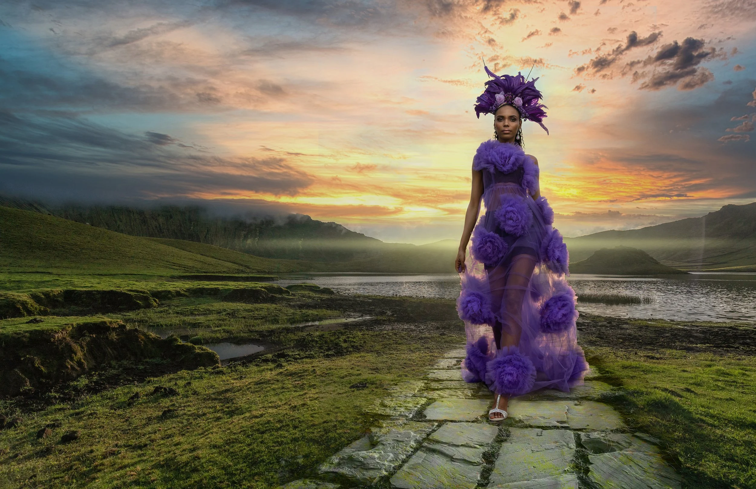 A woman in a purple, elaborate dress with large purple pom-poms, walking on a stone path across a river, with a sunset over a mountainous landscape in the background.
