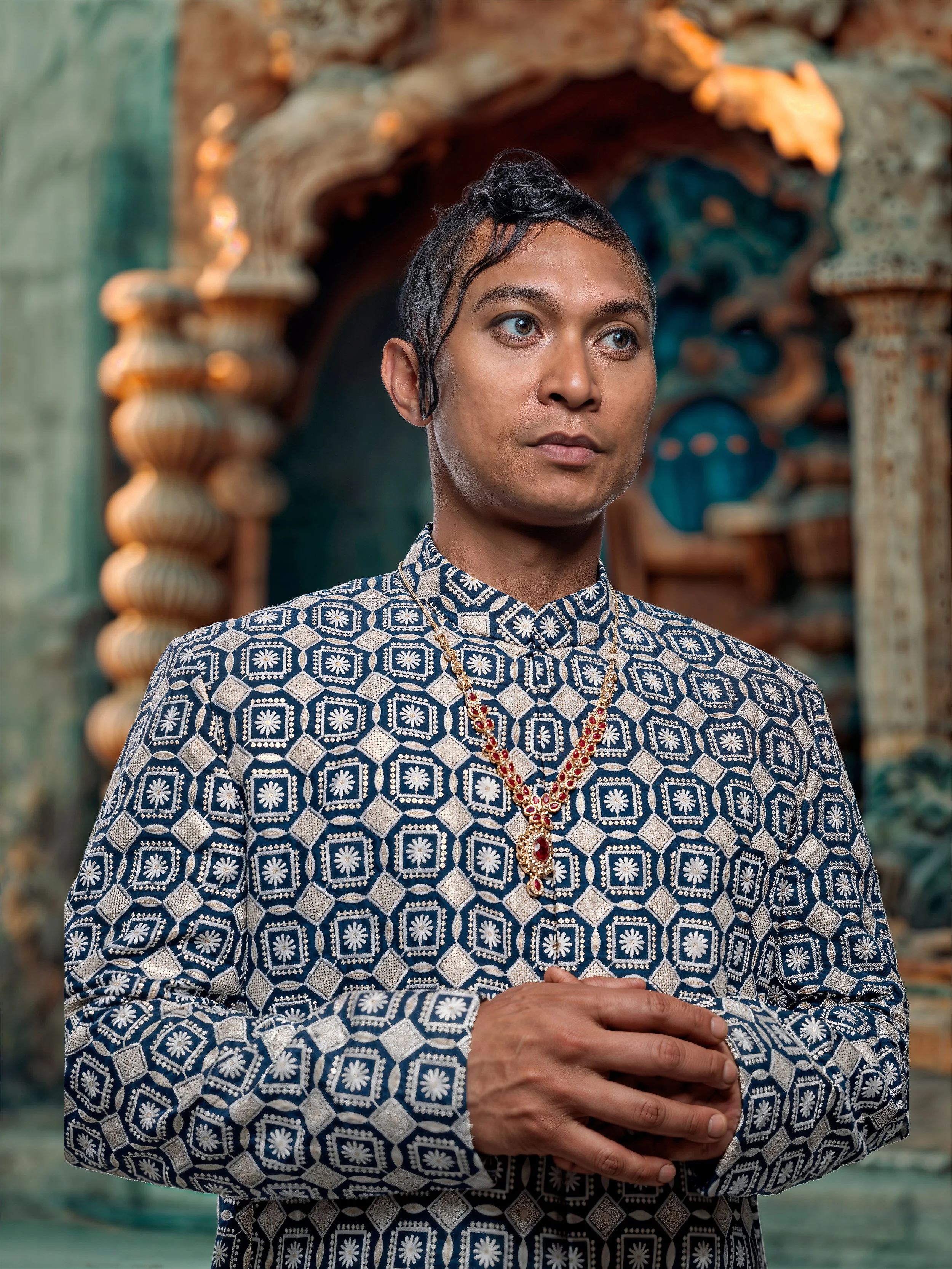 A person dressed in traditional Indian attire with jewelry, standing in front of an ornate, decorative background.