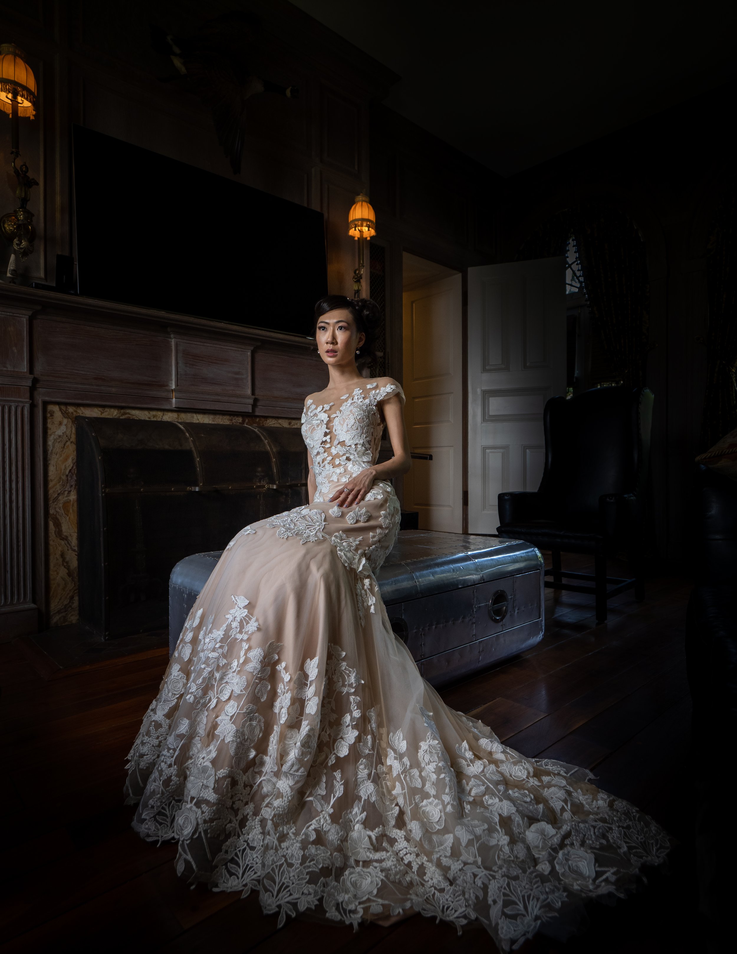 A woman in an elegant, lace wedding dress sitting on a metallic chest in a dimly lit, vintage-style room with dark wood paneling and furniture.