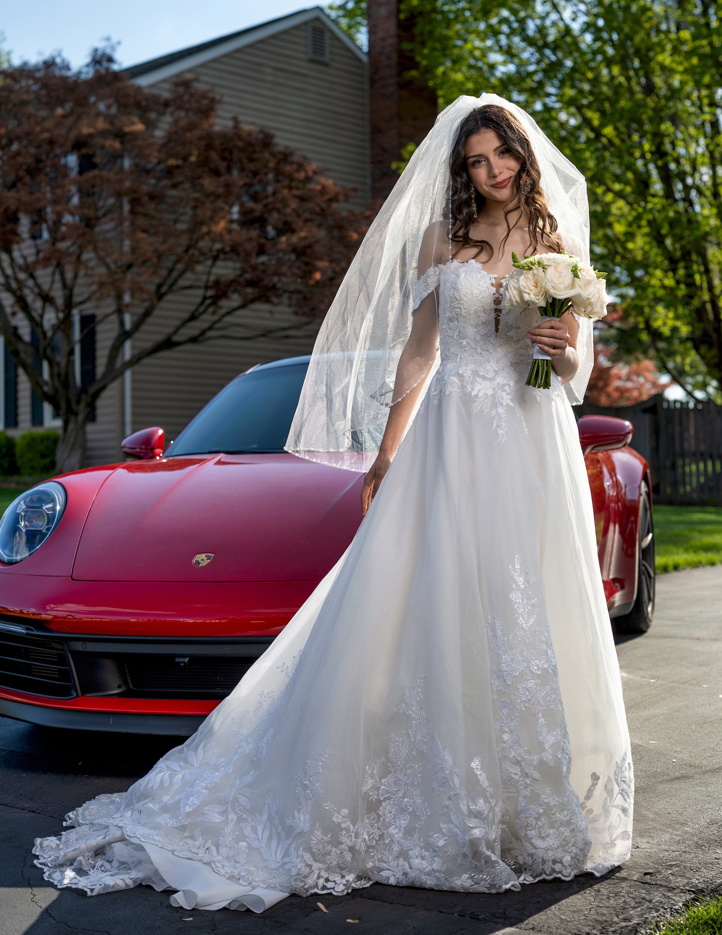 A bride in a white wedding dress and veil holding a bouquet of white roses, standing outdoors in front of a red sports car with a house and trees in the background.