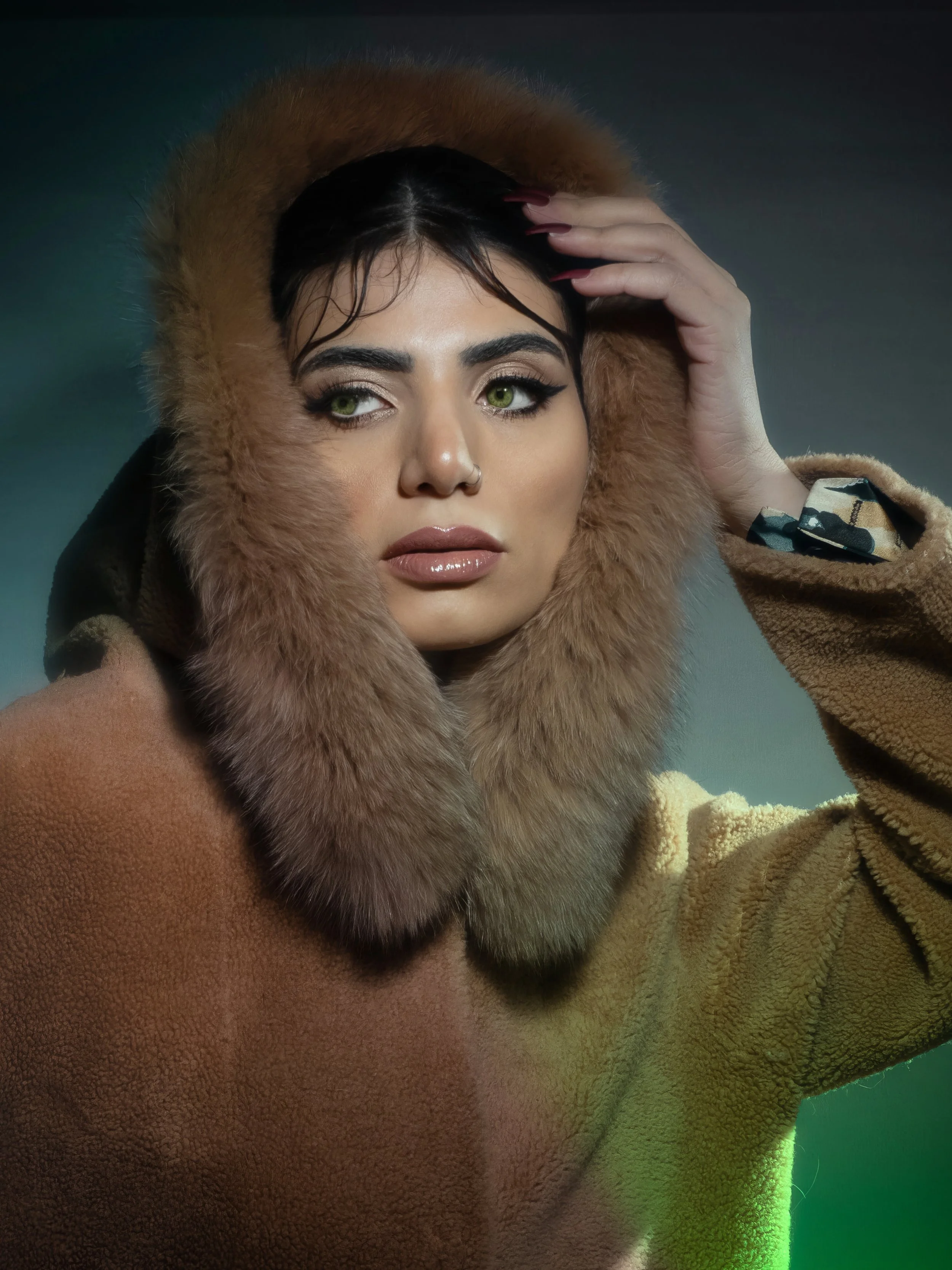 A woman with dark hair, green eyes, and full lips wearing a brown furry hooded coat, looking off to the side.