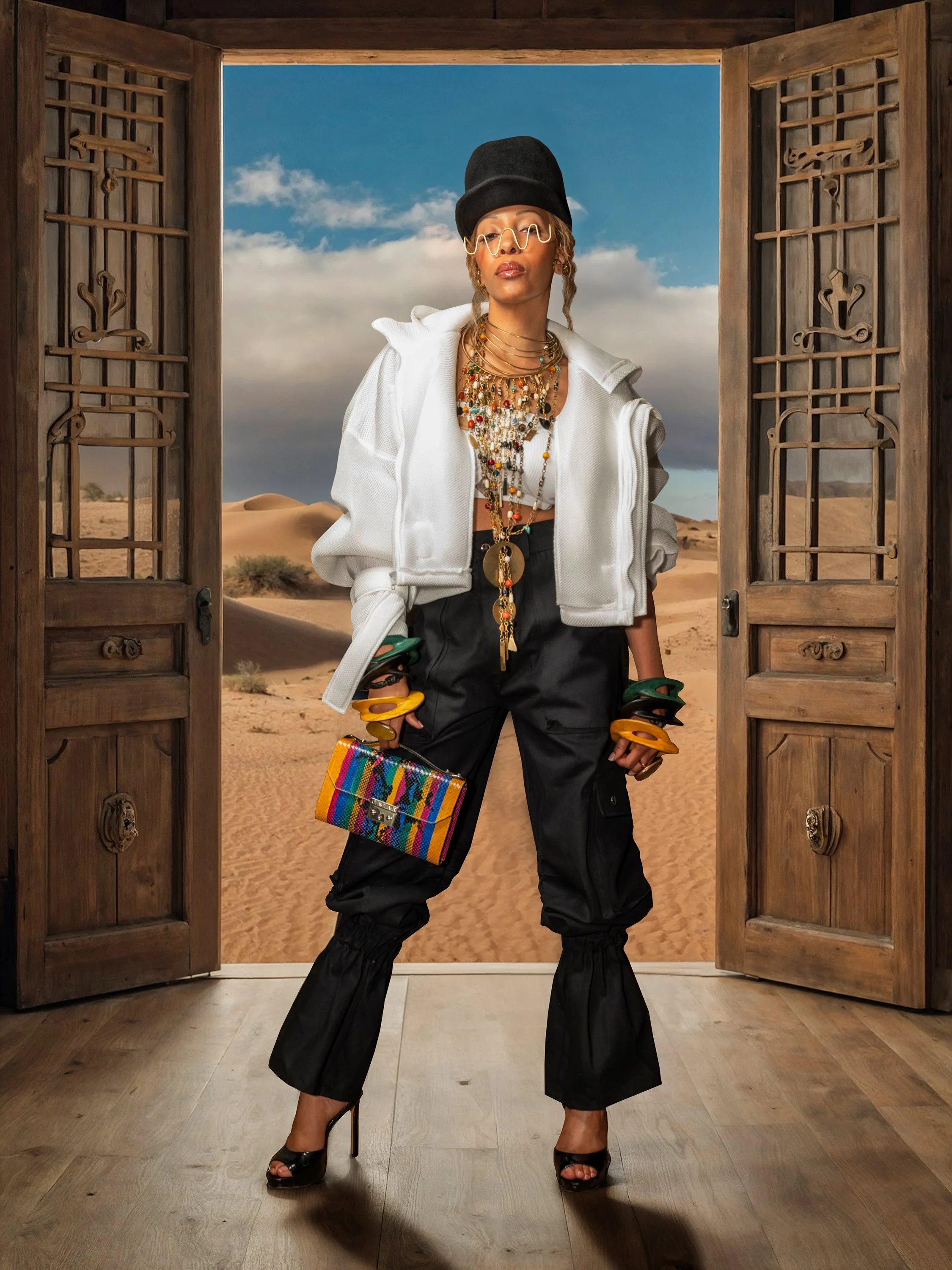 A woman standing in a room with open wooden doors, desert landscape with sand dunes behind her, wearing stylish clothing and jewelry, holding colorful bangles and a small multicolored bag.