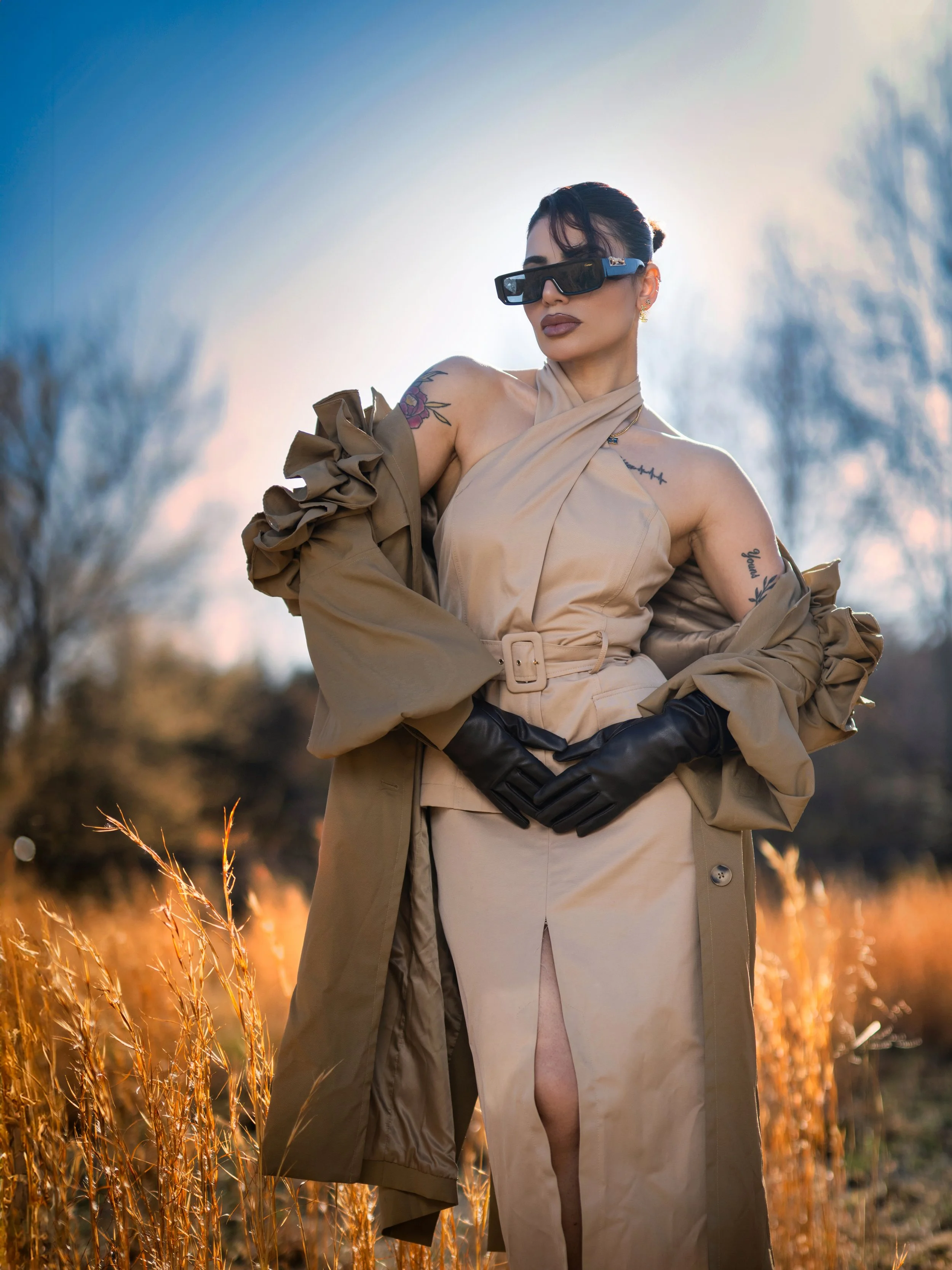 A woman standing in a field with tall golden grass during sunset, wearing sunglasses, a beige dress with a slit, black gloves, and a brown trench coat draped over her shoulders.