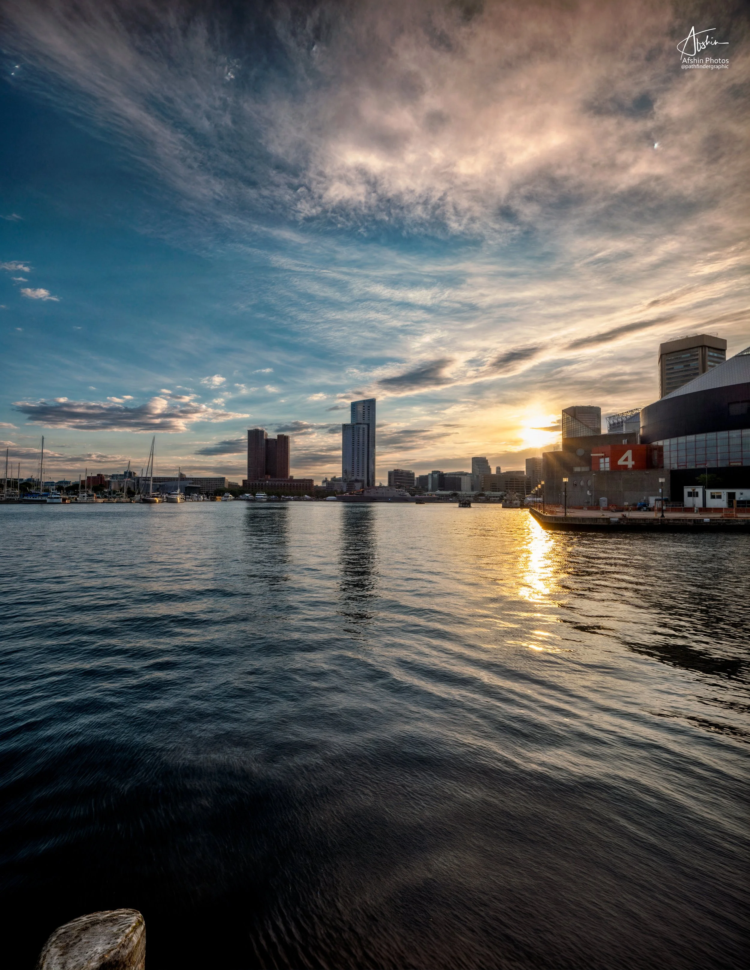 Sunset over a city skyline with water in the foreground, displaying river, boats, and skyscrapers.