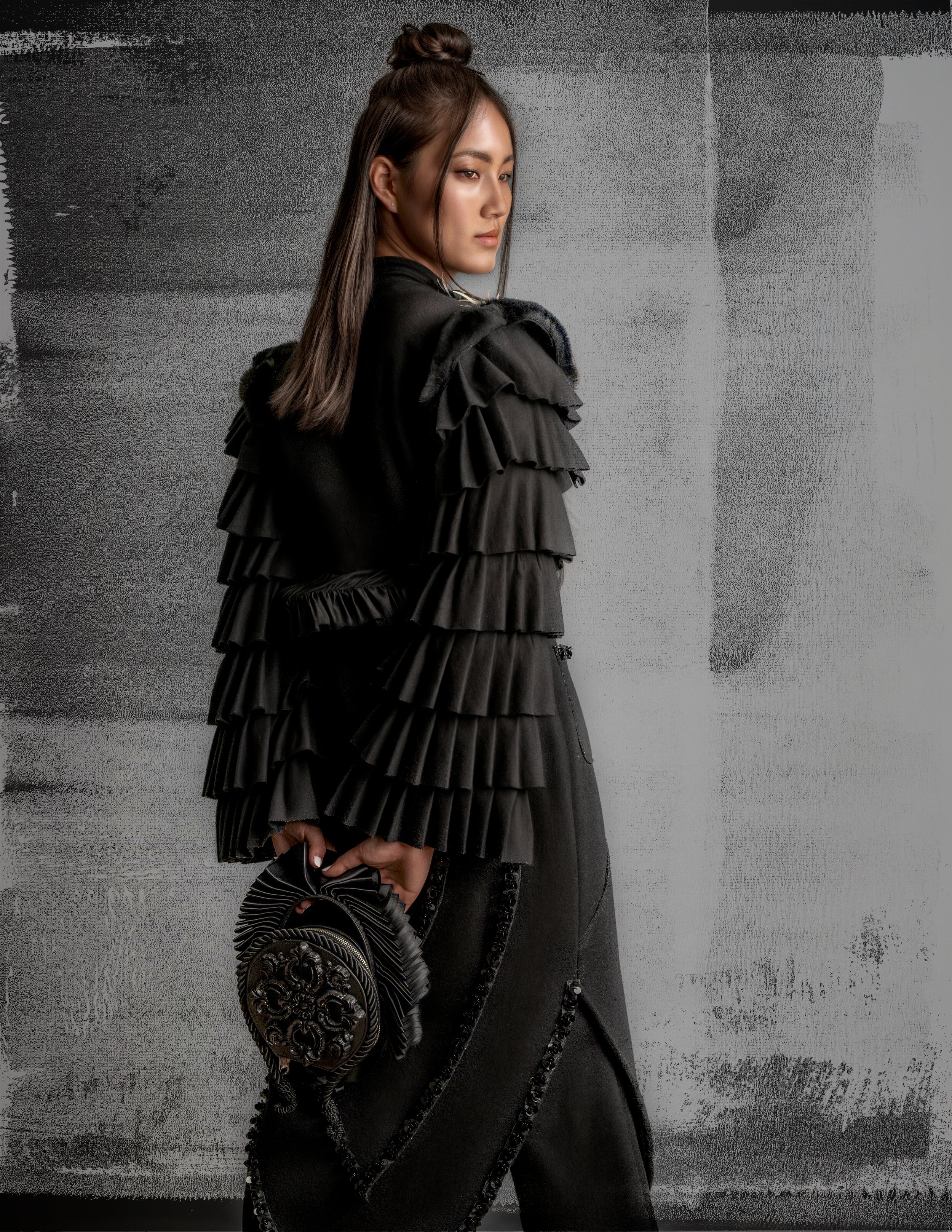 A woman dressed in a black outfit with tiered ruffle sleeves and a long skirt, holding a round black handbag with a decorative clasp, standing against a textured gray background.